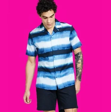 Young man with dark curly hair wearing a blue and white striped short-sleeve button-up shirt and dark shorts, standing against a pink background.