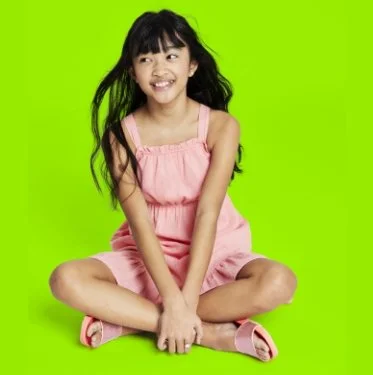 Young girl with long black hair, wearing a pink dress, sitting cross-legged on a green background, smiling and looking to the side.
