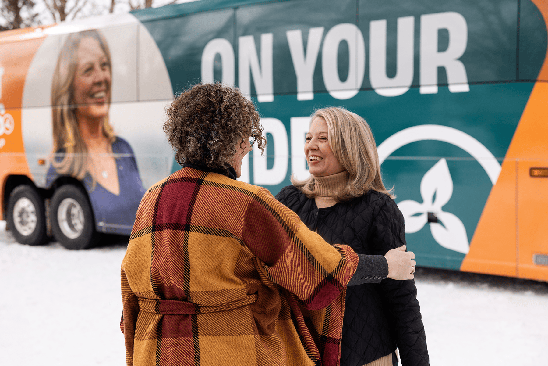 Ontario NDP leader Marit Stiles speaking with a supporter beside a campaign bus branded with “On Your Side.”