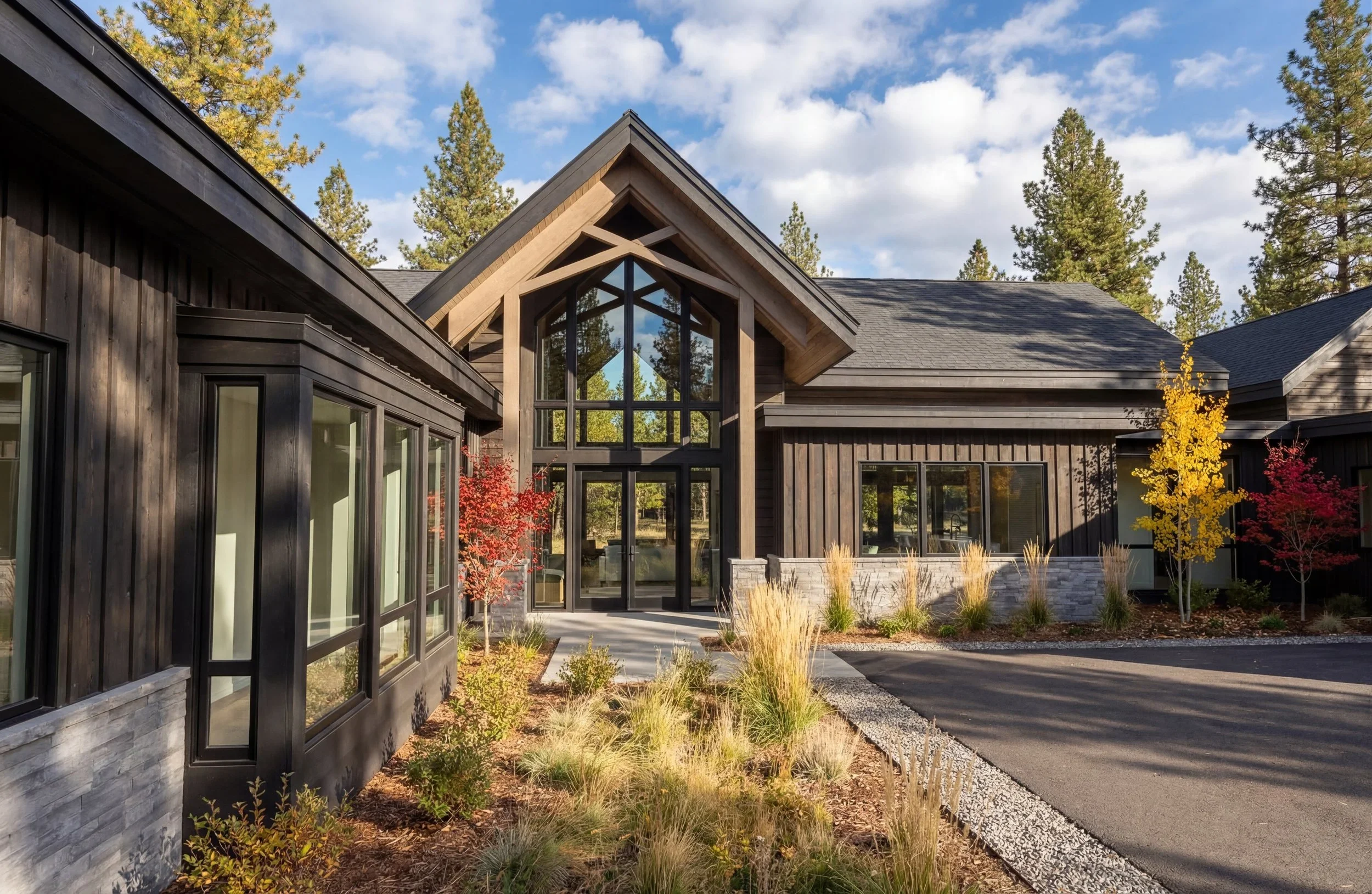 Front view of a modern house with black wooden exterior, large glass entryway, surrounded by trees and autumn-colored shrubs, under a partly cloudy sky.