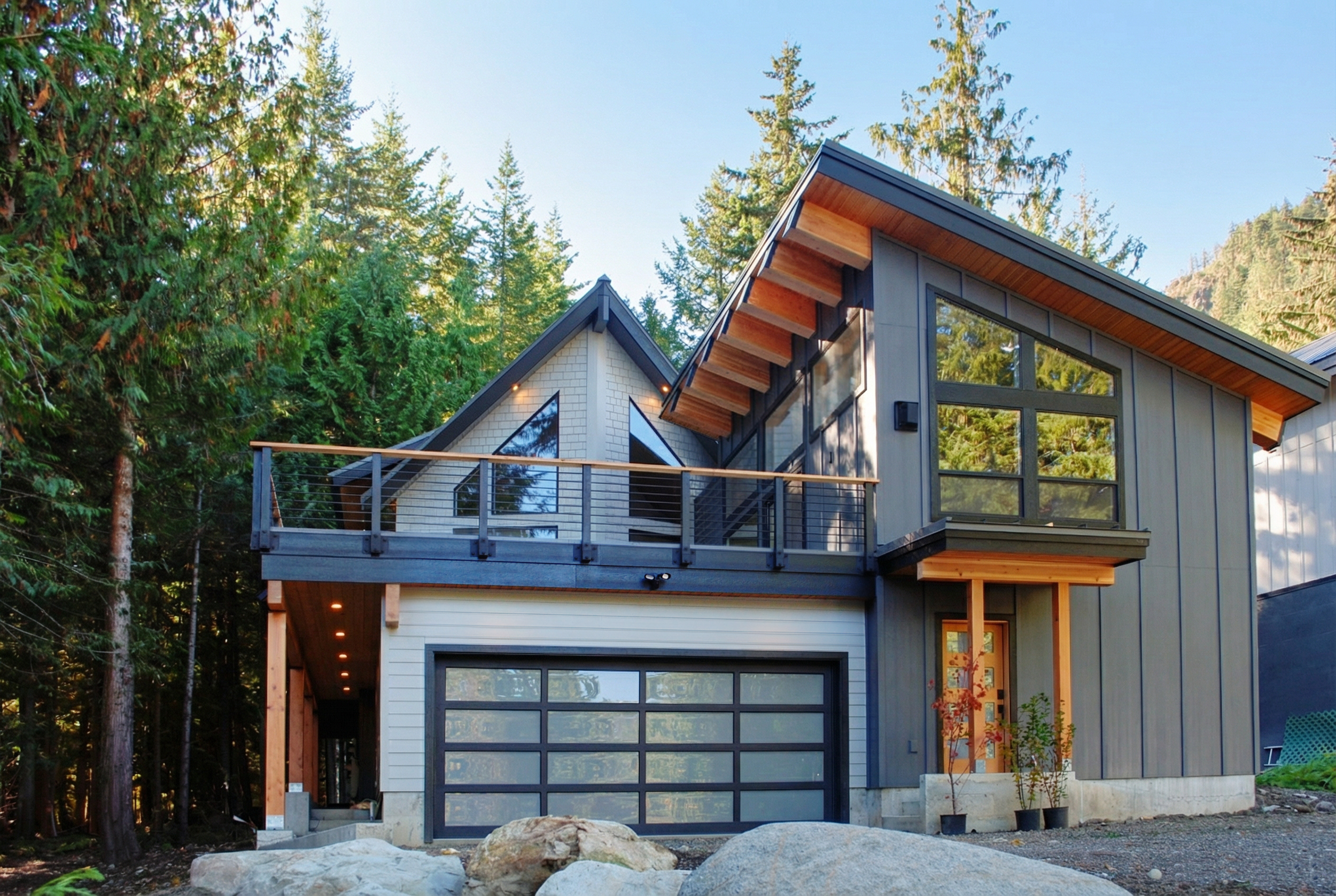 Modern house with large windows, a balcony, and a garage, surrounded by tall pine trees.