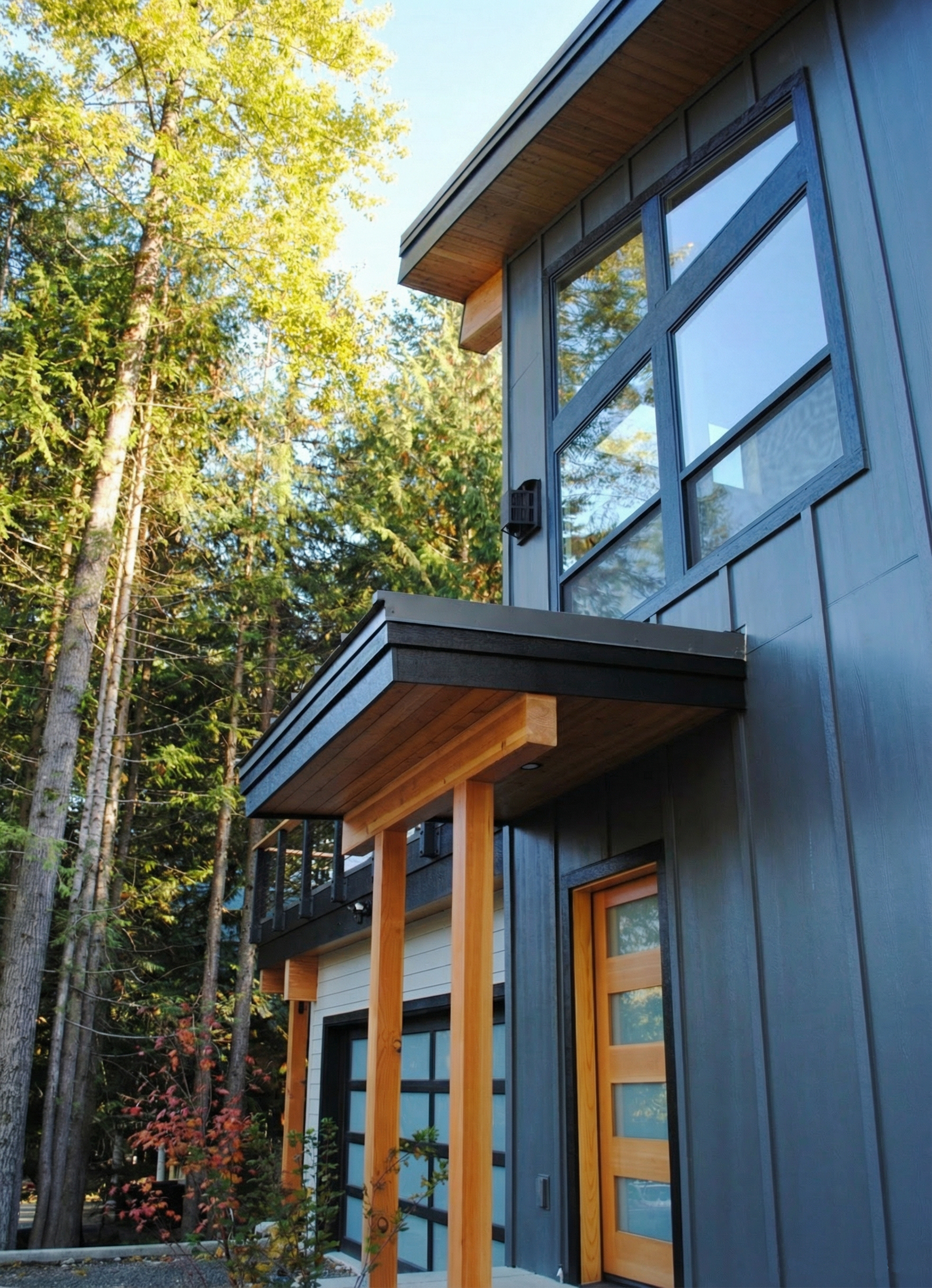 Modern house with dark gray exterior siding, large glass windows, and a small balcony with a black railing, surrounded by trees with green foliage.