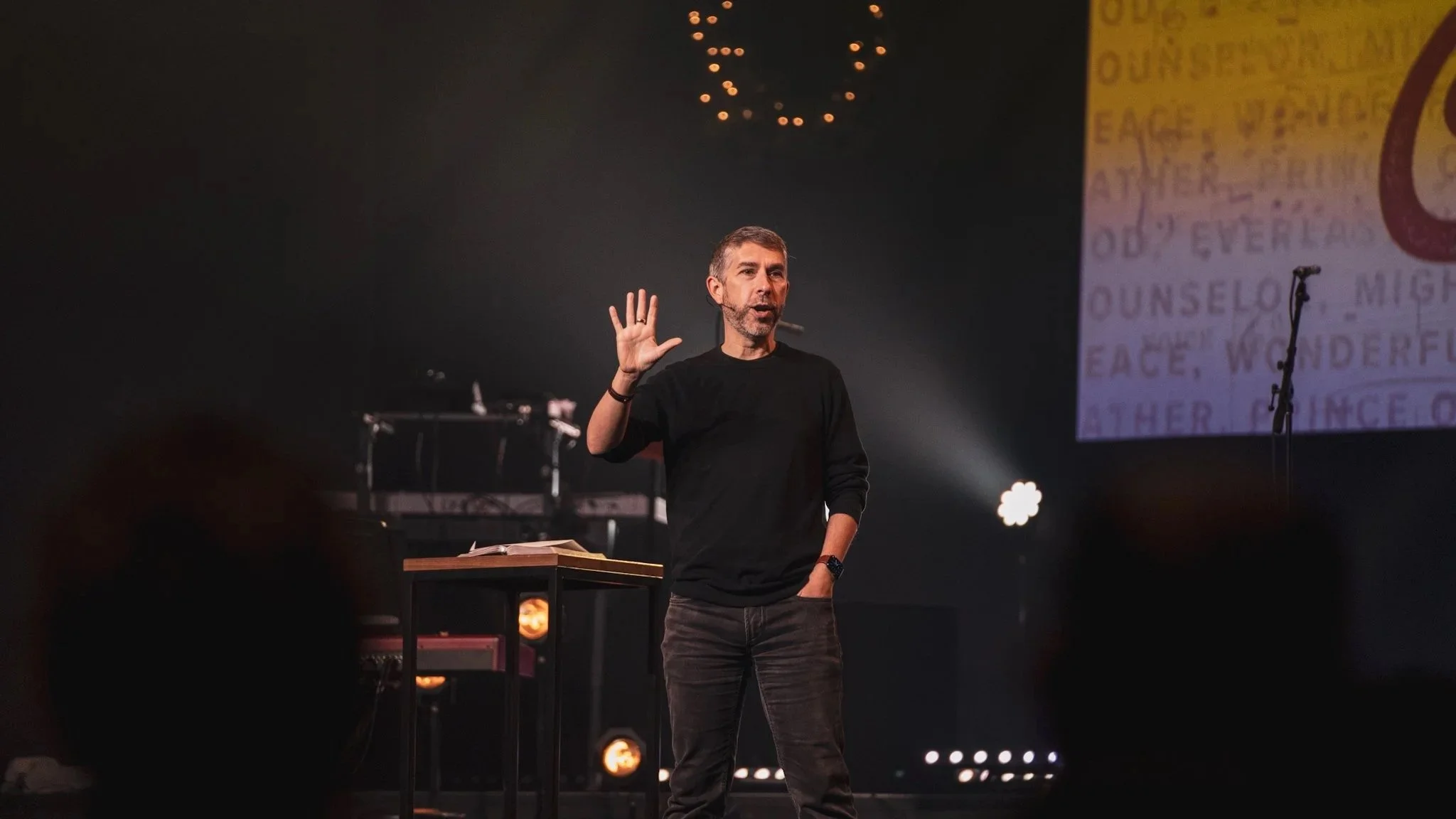 Speaker Brad D. Jenkins, with short gray hair and beard, delivering a presentation on stage at Anthem Church with a dark background, illuminated by stage lights, with a large screen displaying text behind him.