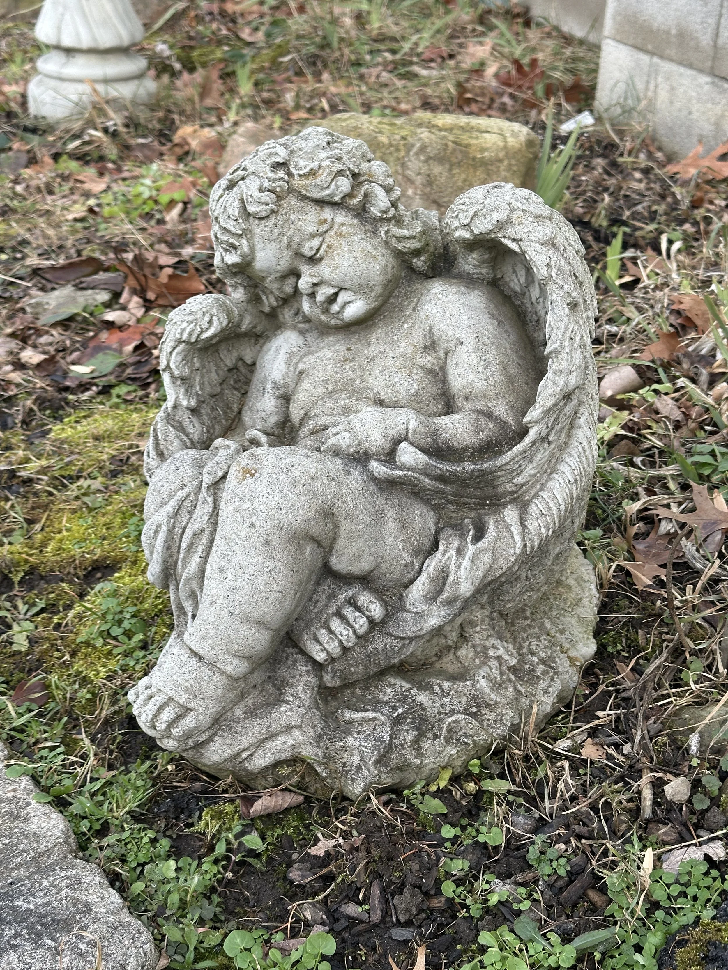 A stone statue of a young angel with curly hair, wings, and a peaceful expression, holding an open book, placed in a garden area with soil, moss, and fallen leaves.