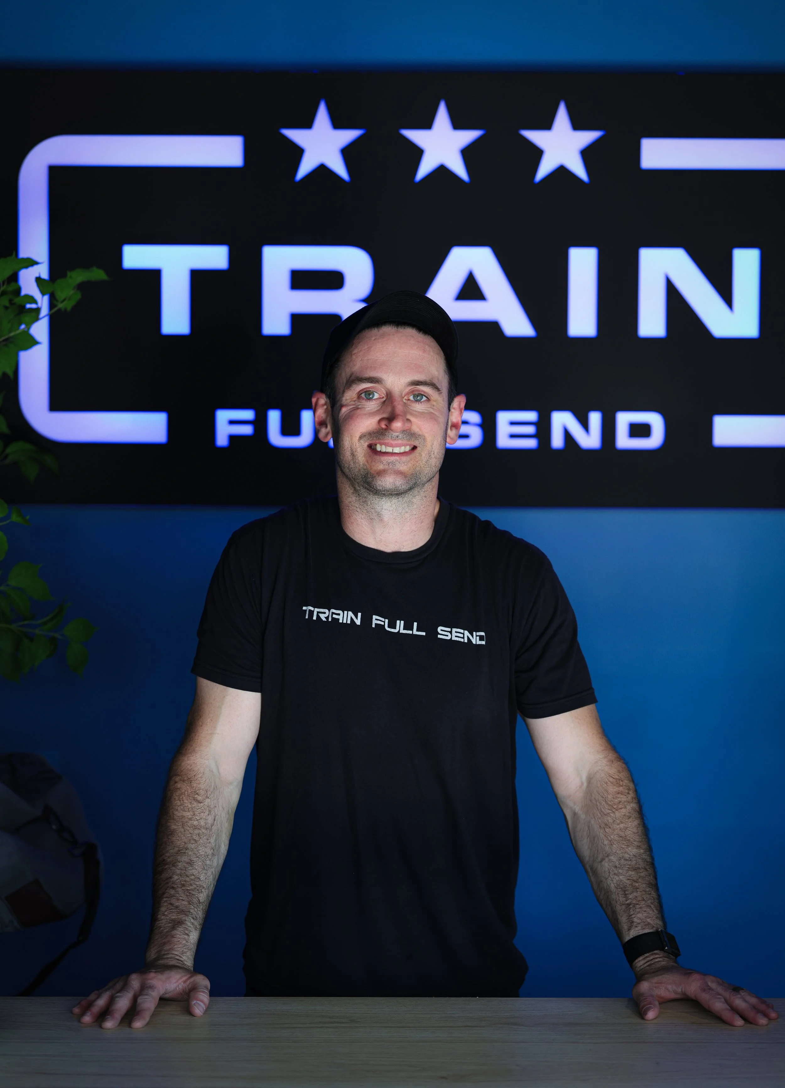 A smiling man with short dark hair and light facial hair standing with arms crossed inside a gym, wearing a blue t-shirt with 'PEX' logo and a fitness tracker on his wrist, with gym equipment behind him.