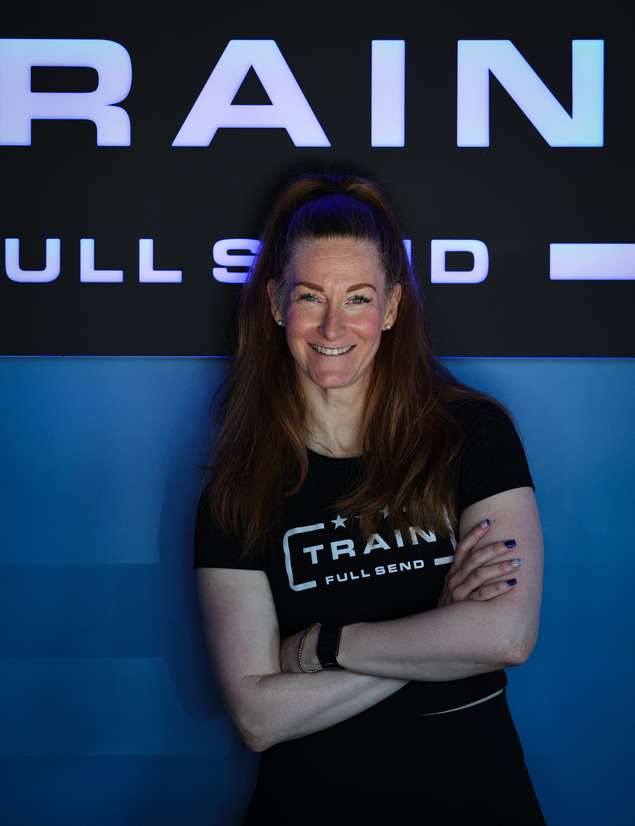 A woman with red hair tied in a high ponytail, wearing a black long-sleeve athletic top, smiling and standing with arms crossed against a gray textured wall.