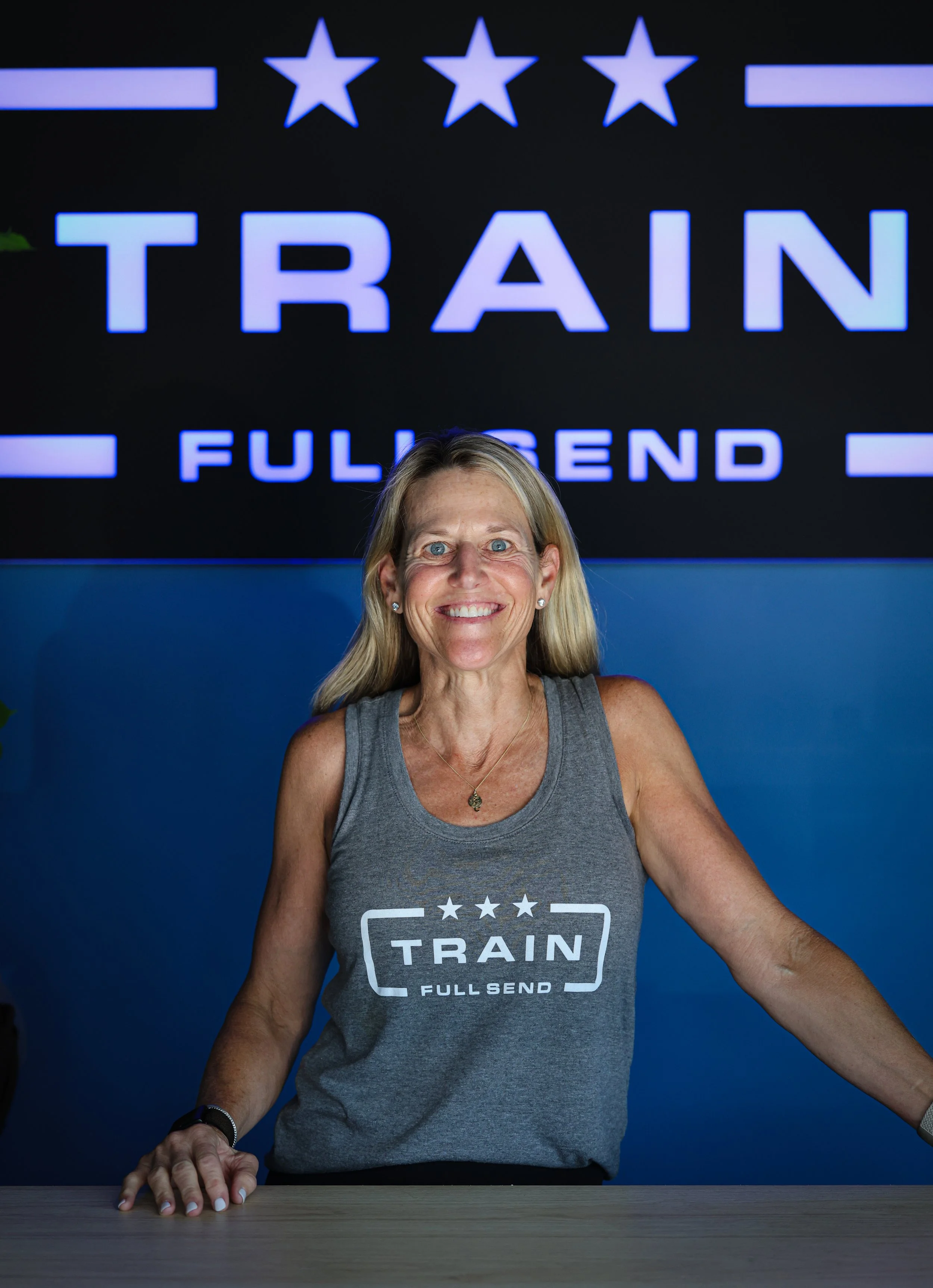 A tattooed woman wearing a white cap and gray tank top with the word 'REFUGE' on it, raising her right arm in a gym-like setting with a dark green curtain background.