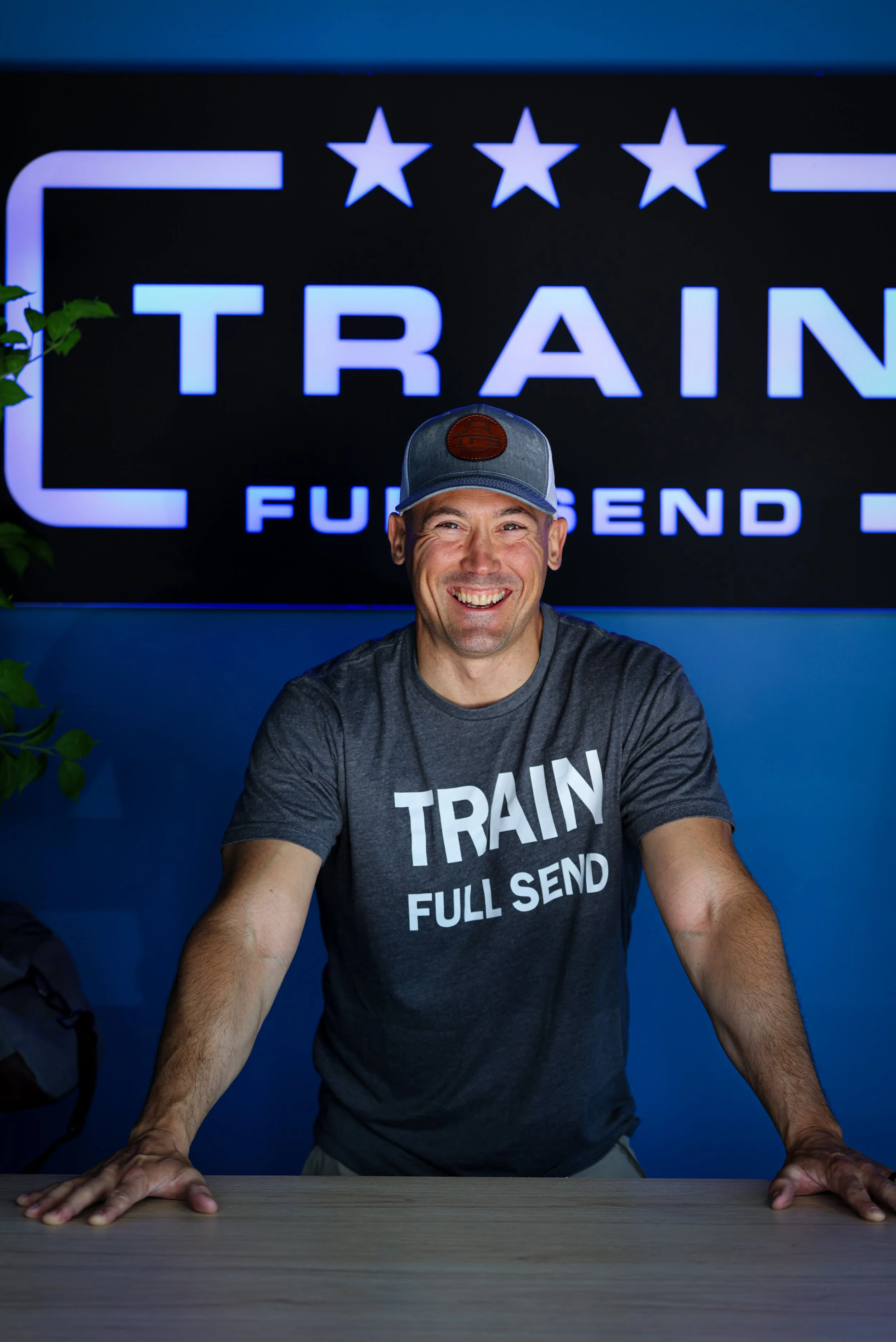 A man standing against a gray wall, wearing a light gray cap with a leather patch and a black t-shirt with white text that says 'TRAIN FULL SEND'.