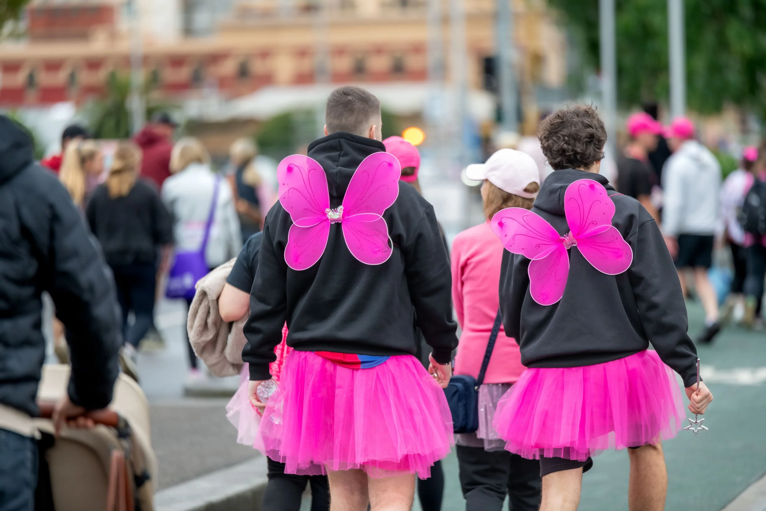 Two racegoers wearing pink fairy wings on their back face away from the camera.