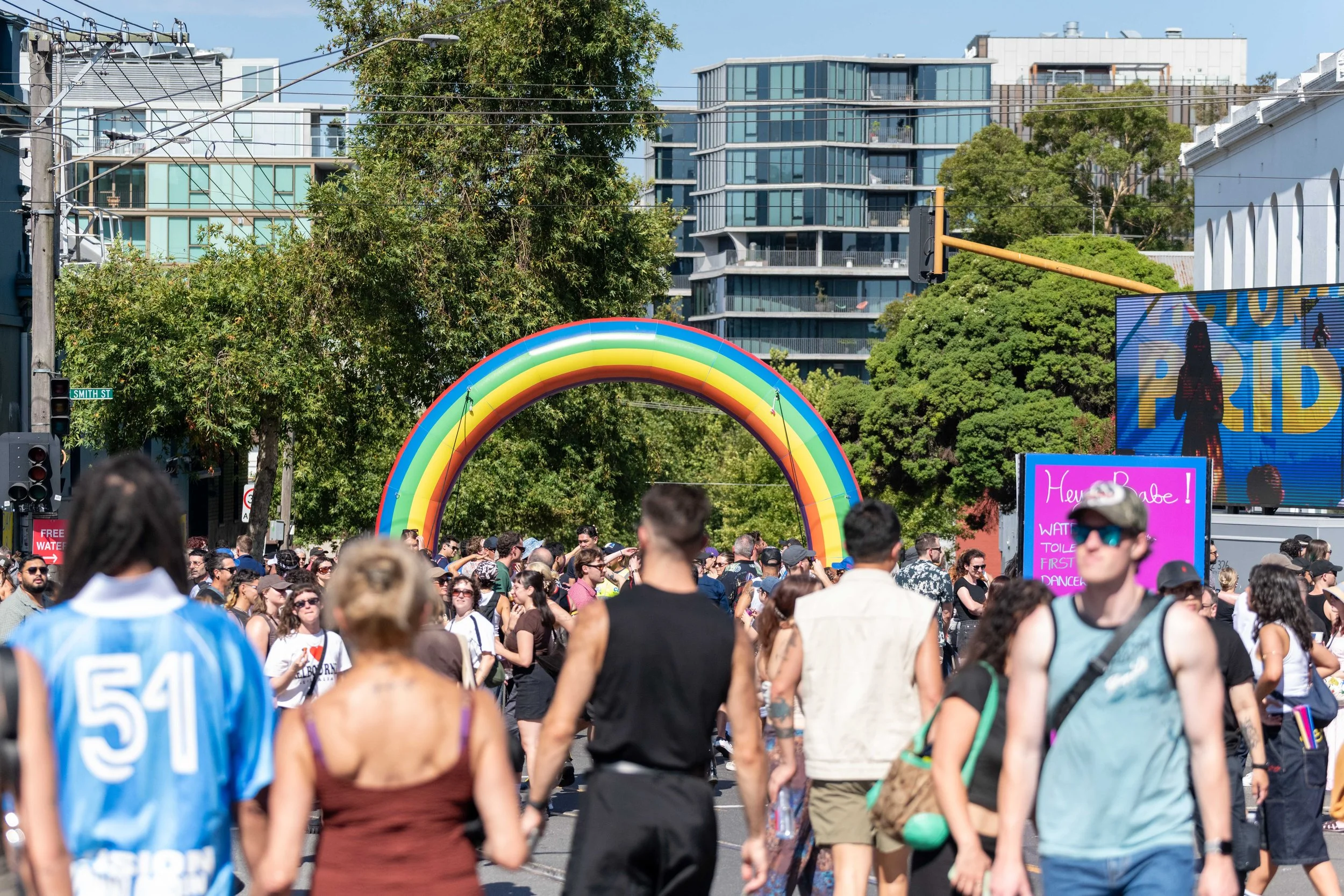 Masses of humans walk along the street. A large inflattable rainbow is seen in the background.