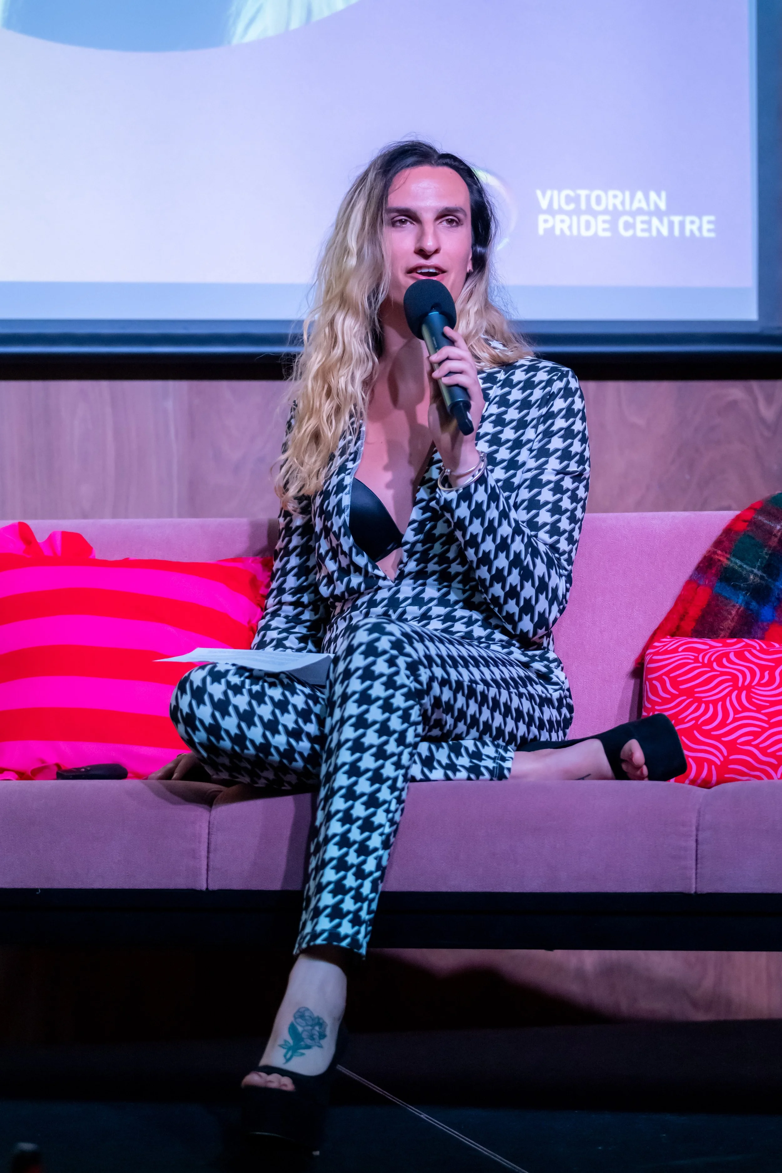 A human in a houndstooth suit sits on a couch and speaks into a microphone.