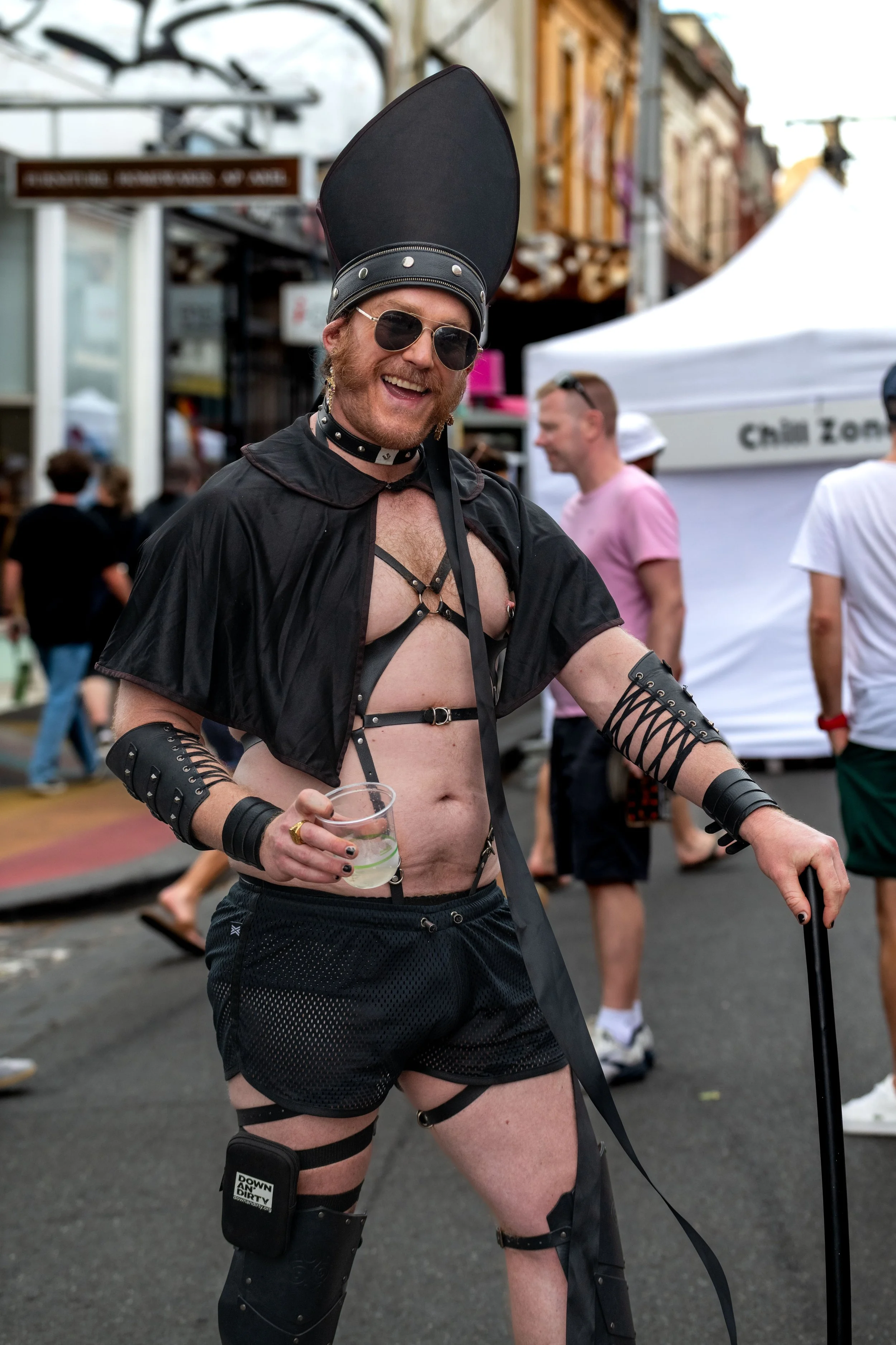Father Daddy wears a black leather harness, cape, and black pontiff hat and poses for the camera.