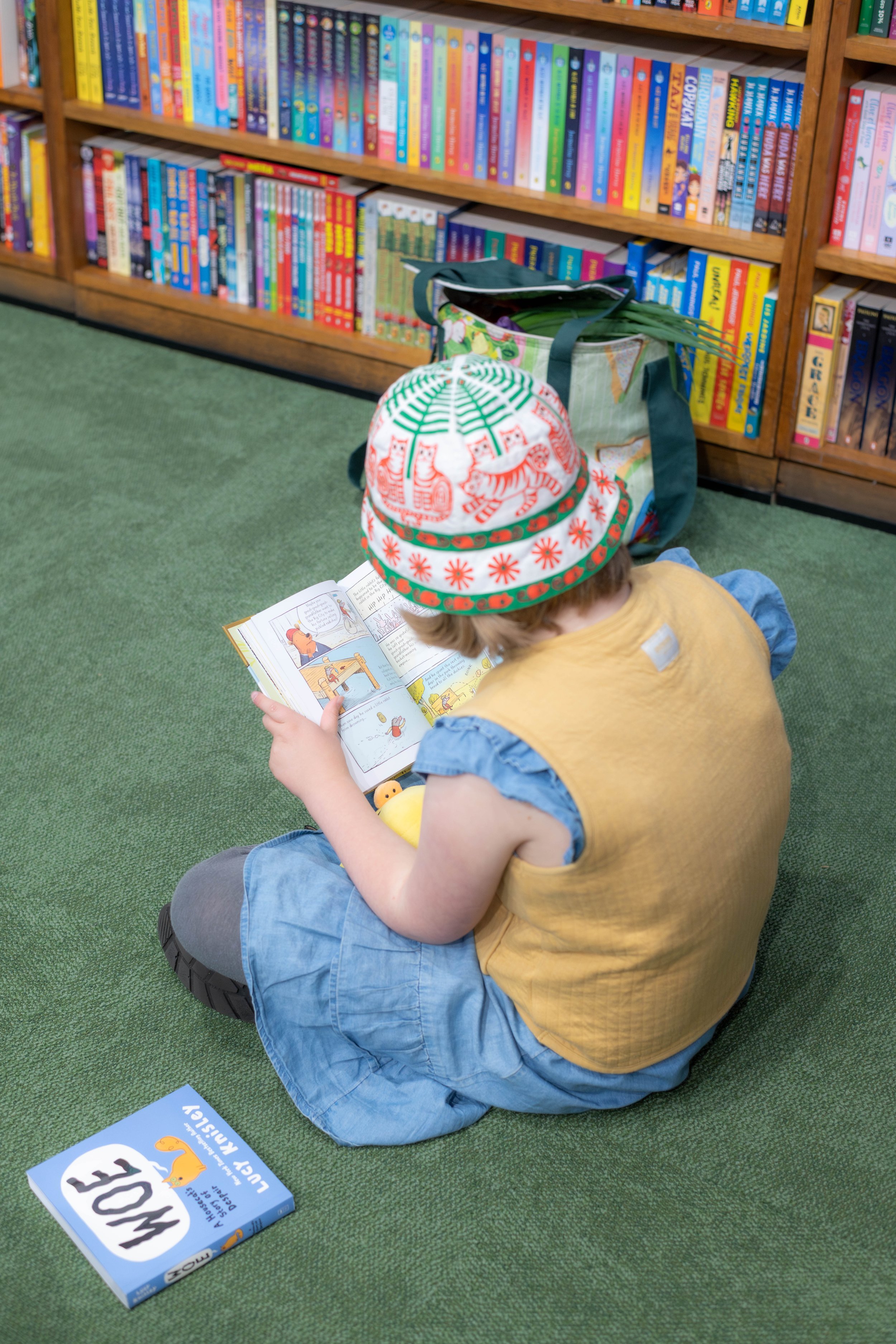 Achild wearing a bucket hat sits cross-legged on the floor whilst reading a book.