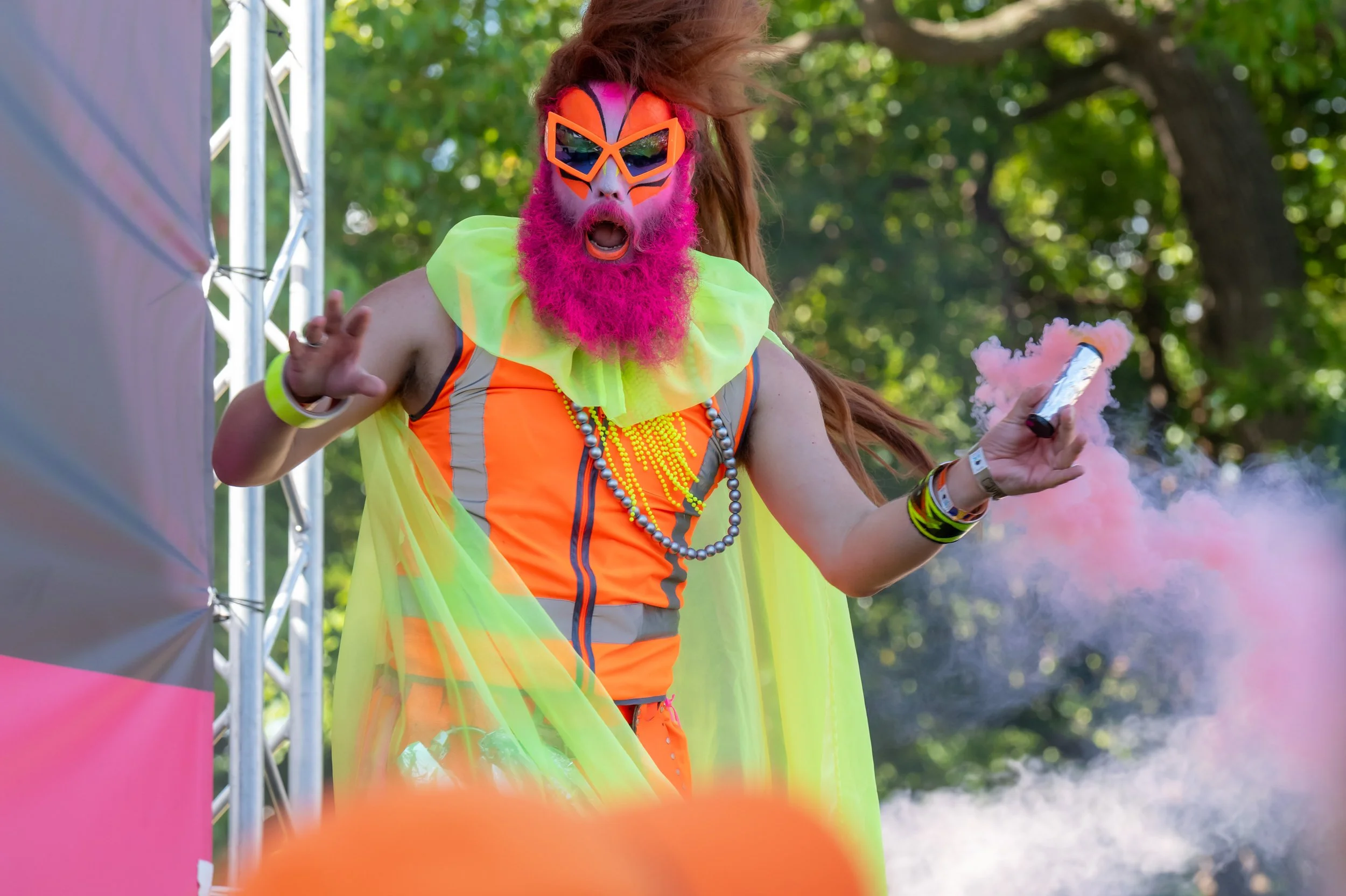 Dandrogyny is dressed in hi-vis clothing and has a pink beard. They are holding a can of pink smoke.