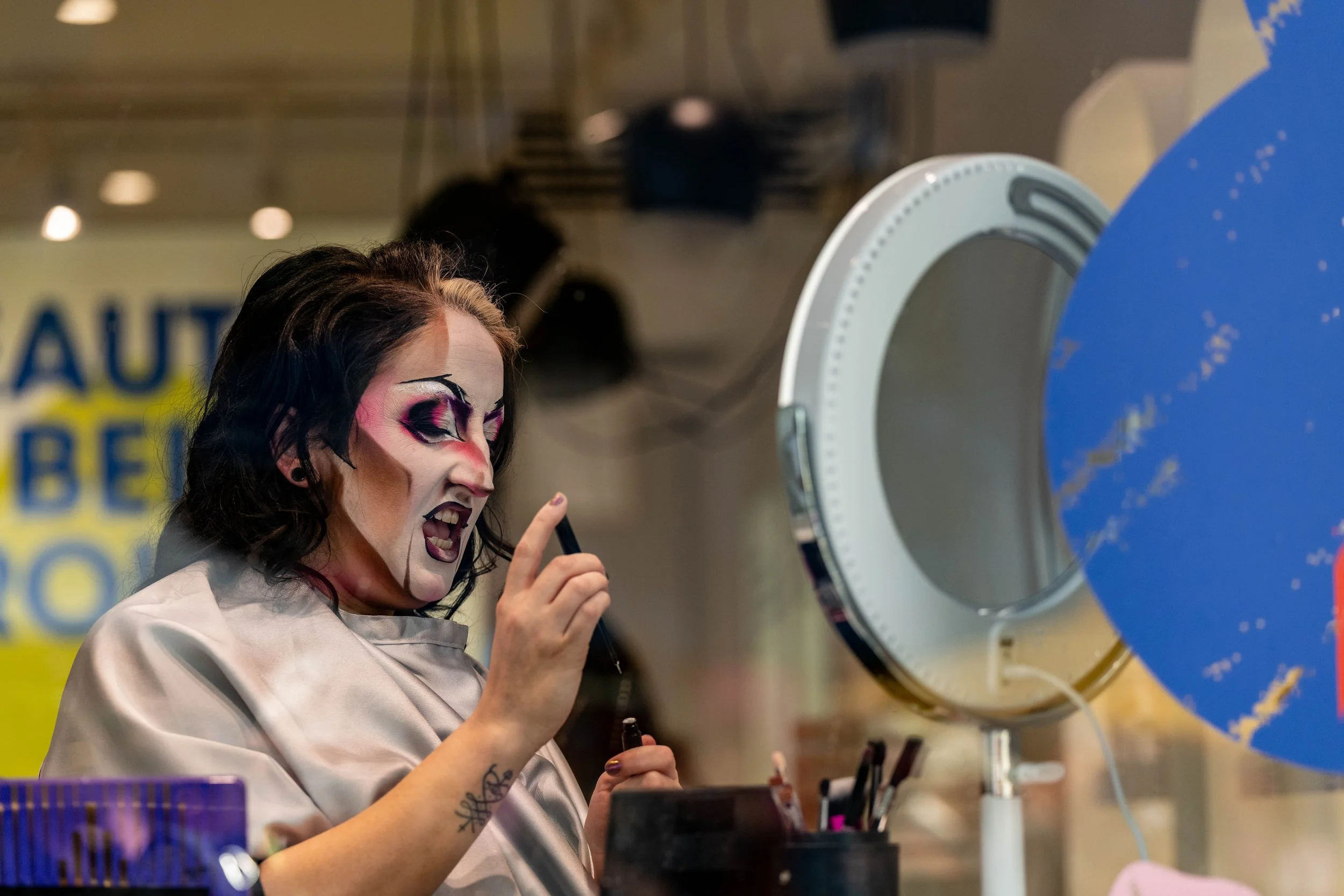 A drag king applies their face paint from behind a glass window.