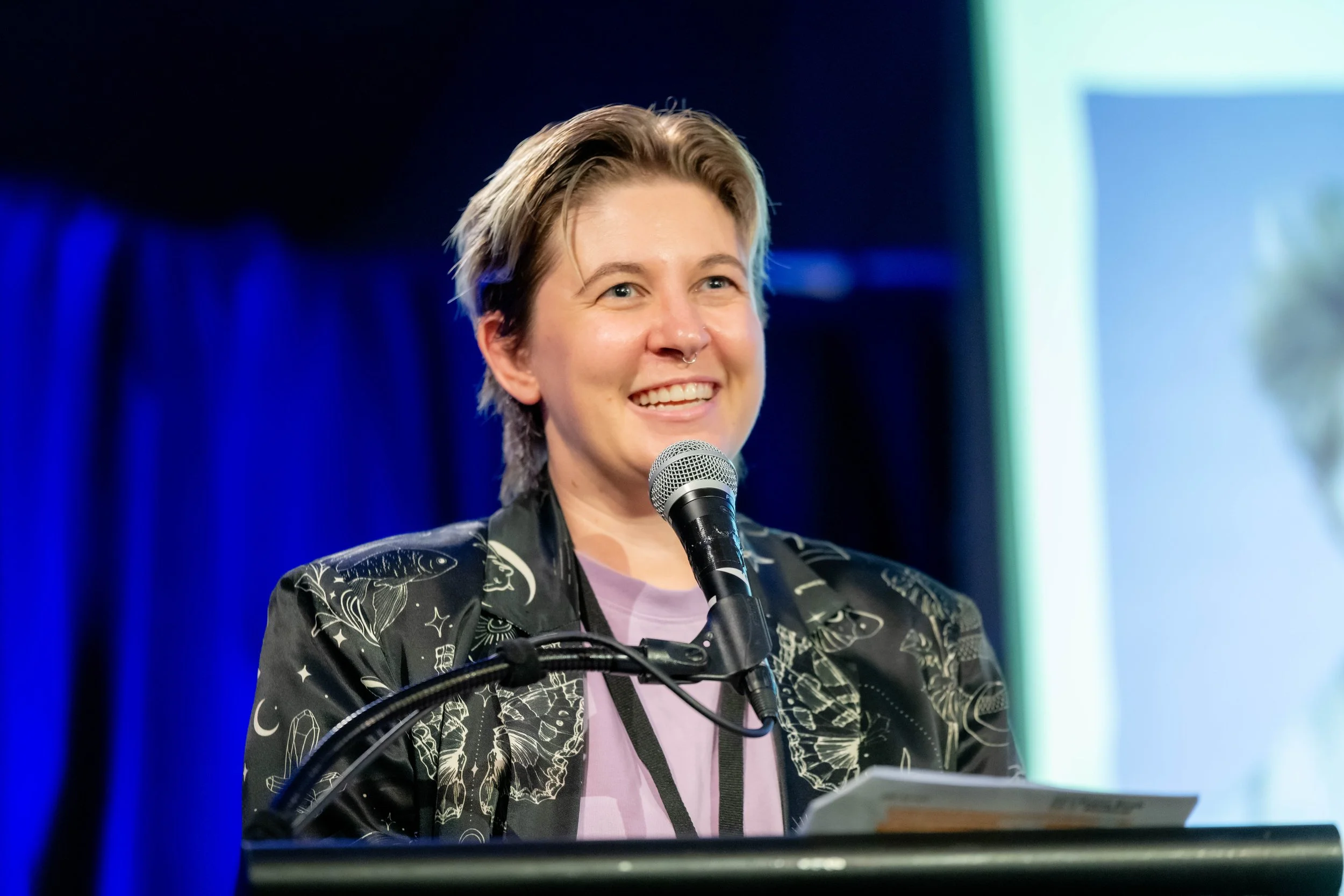 Eli Sutherland wears a black satin jacket whilst standing at a lectern and smiling into a microphone.