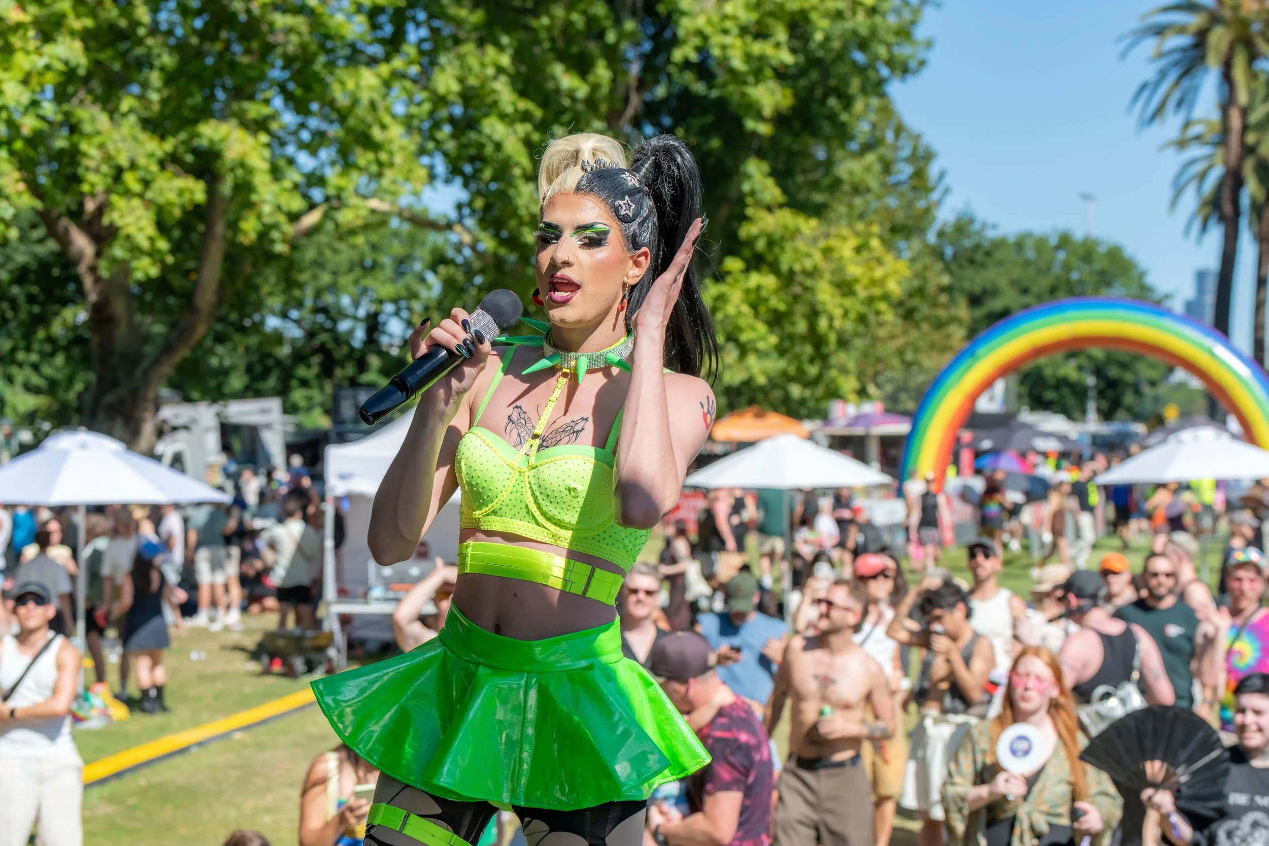 A drag queen wearing green neon performs for the crowd. A large inflatable rainbow is in the background.