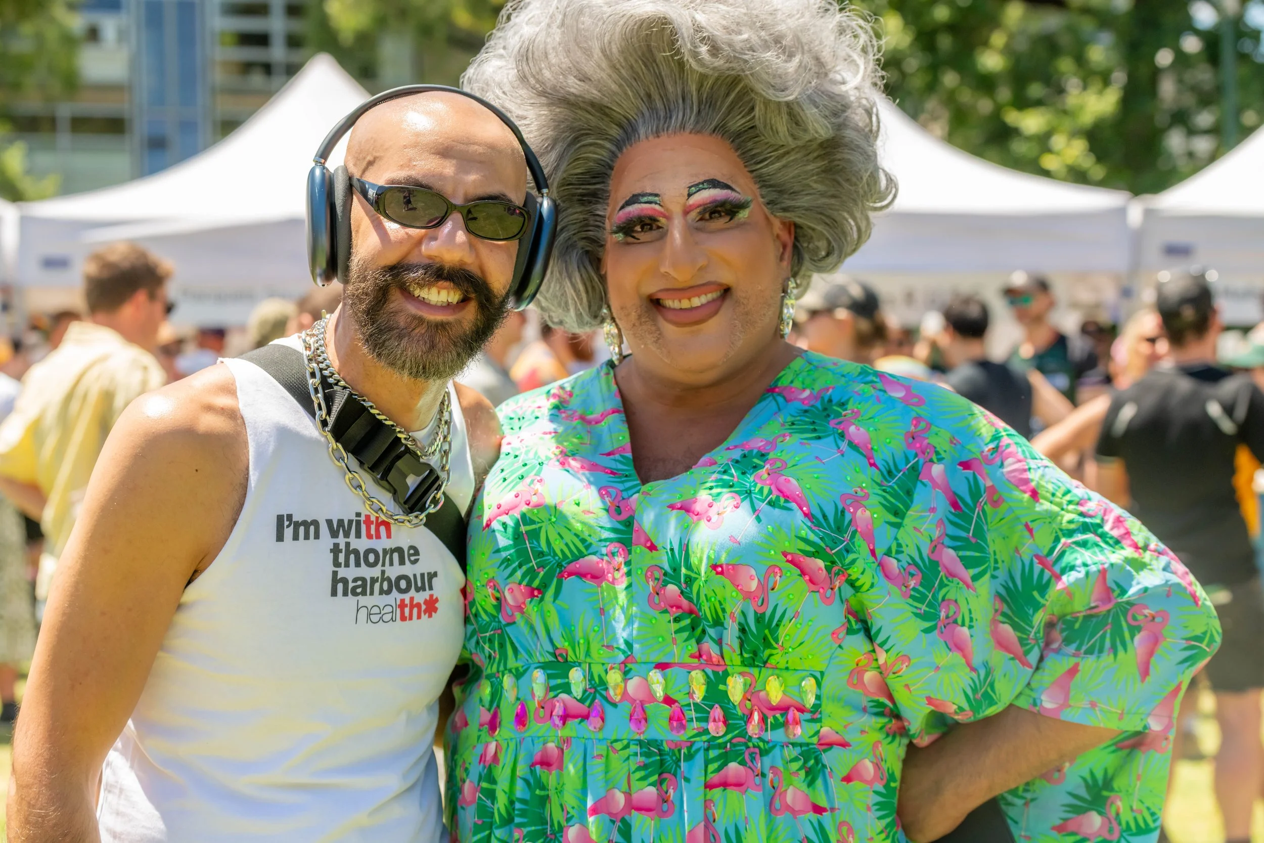 Drag Queen Frock Hudson wears a green pink flamingo mumu and hugs a human wearing ear protectors and a white Thorne Harbour Health singlet.
