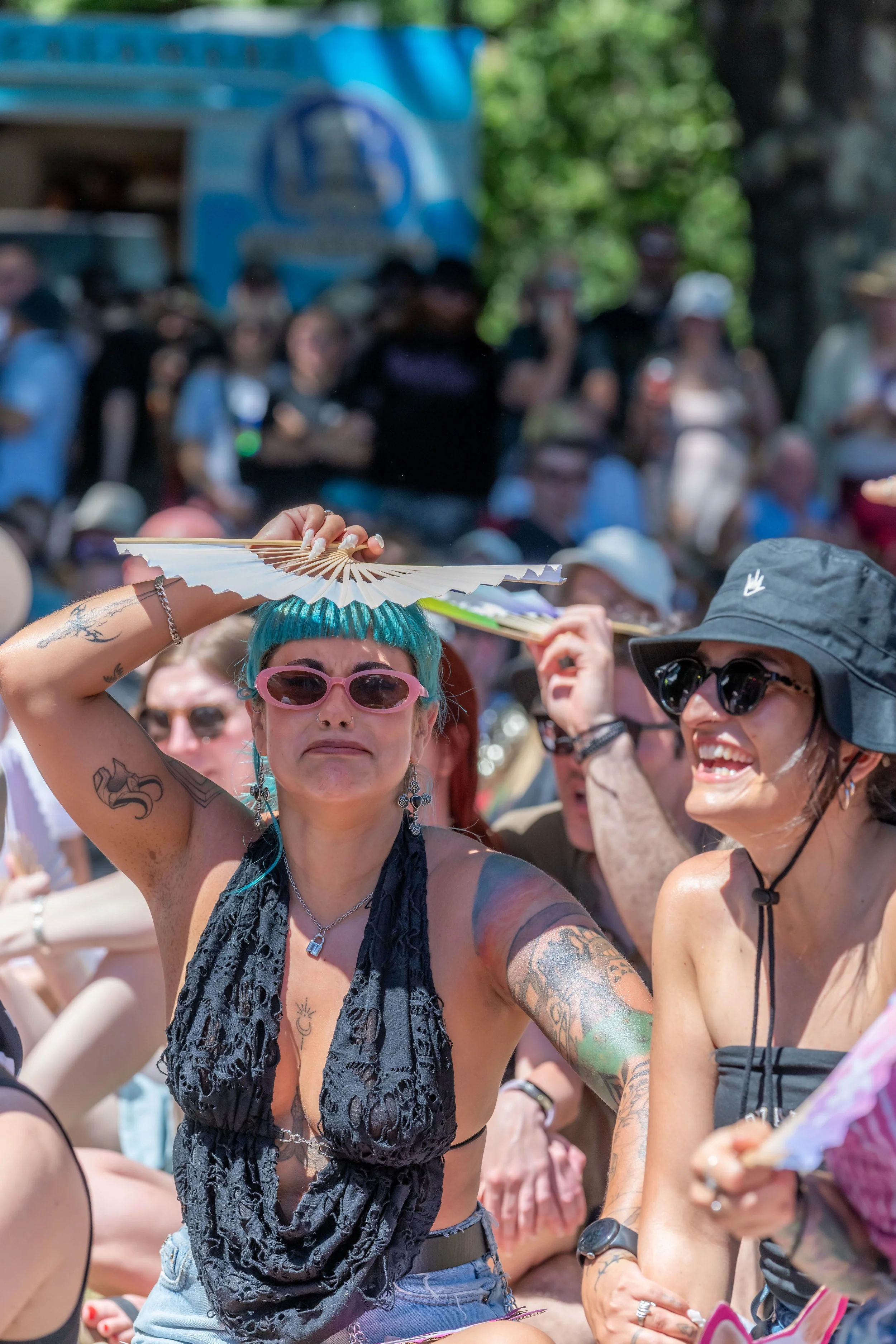 A human wearing pink sunglasses shades themself with a rainbow fan.