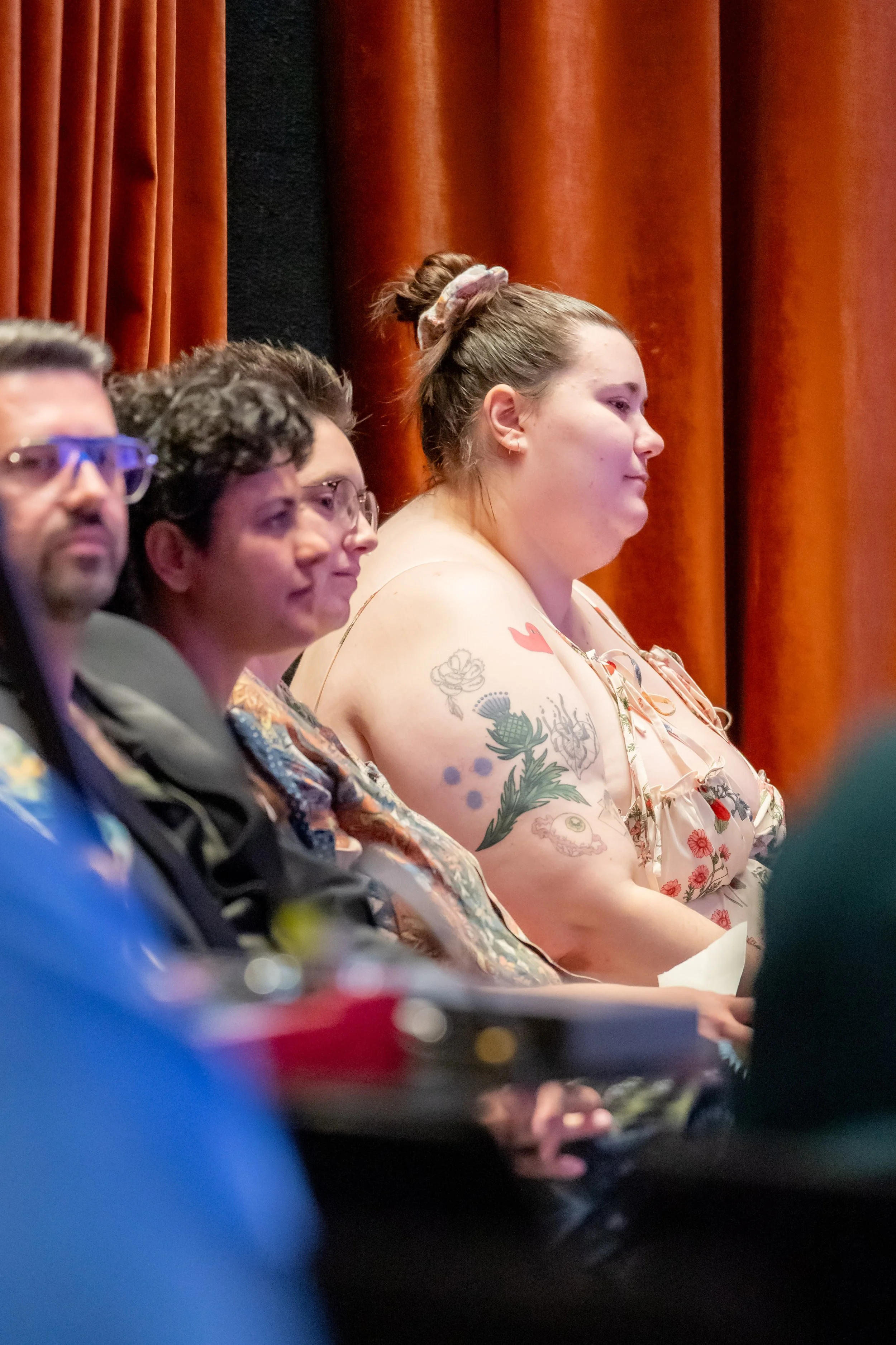 A human with tattoos looks forwards next to other humans looking on.