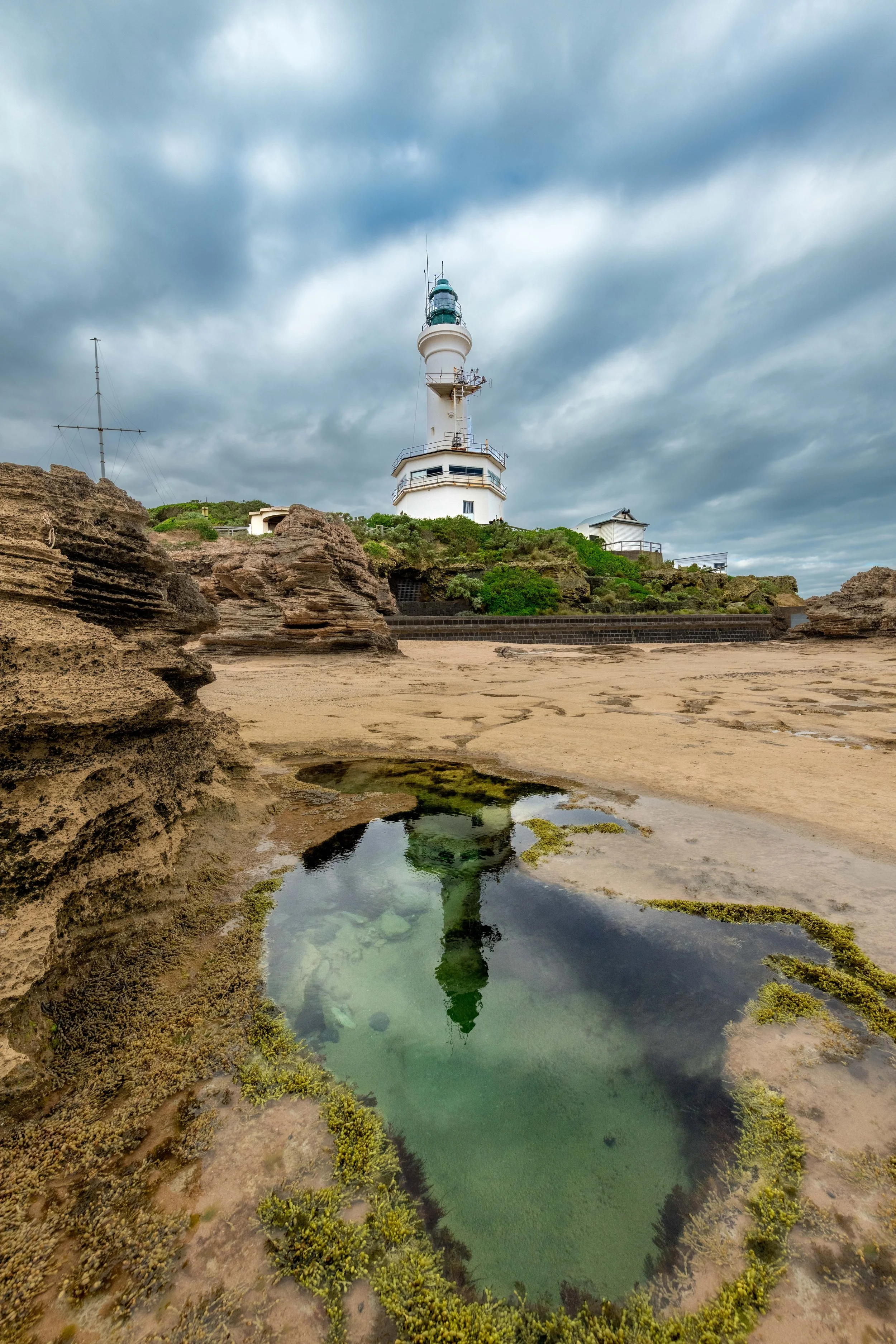 A white lighthouse stands atop a small cliff. Below are rockpools reflecting the lighthouse.
