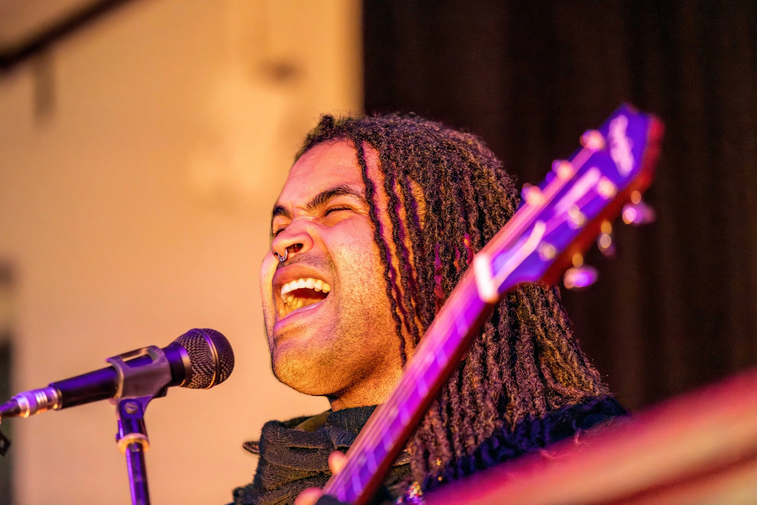 A human holding a guitar sings into the microphone. Their eyes are closed and their mouth is wide open.