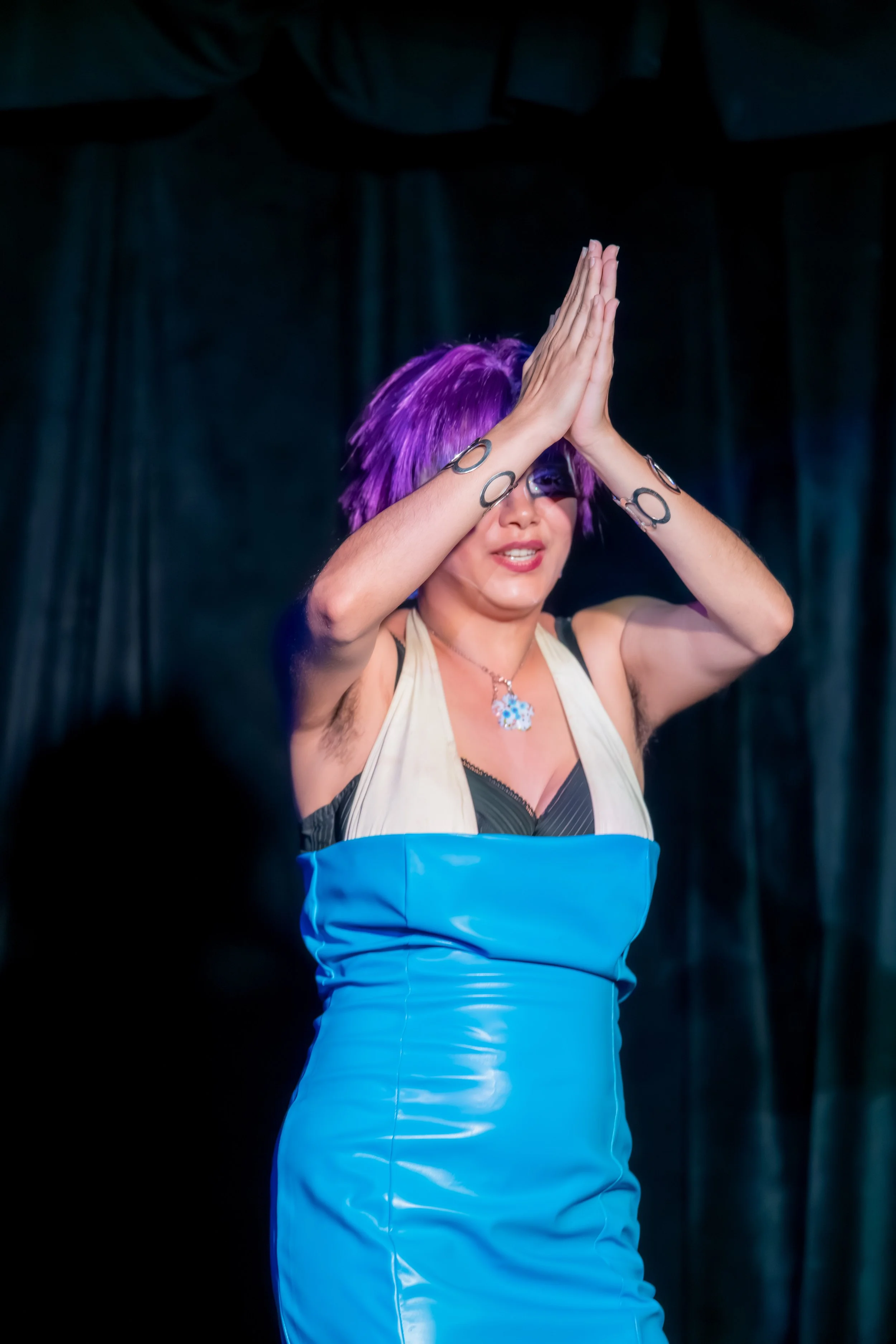A drag queen in a purple wig and blue dress places both palms together in front of their face.