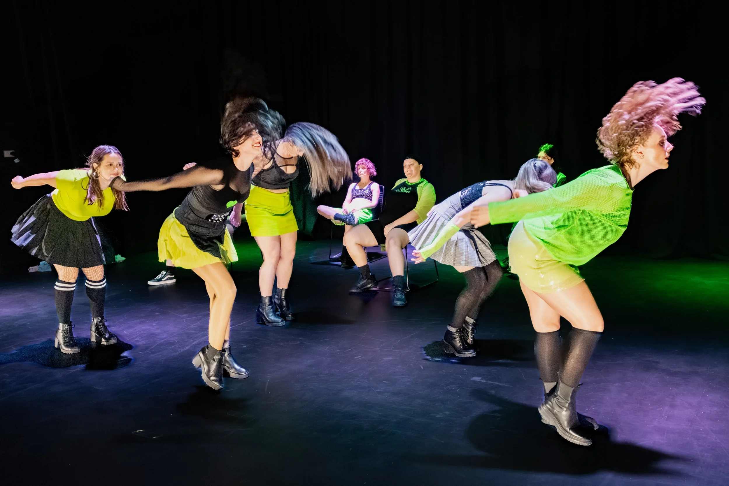 Several dancers in green and black whip their long hair back whilst others sit on chairs.