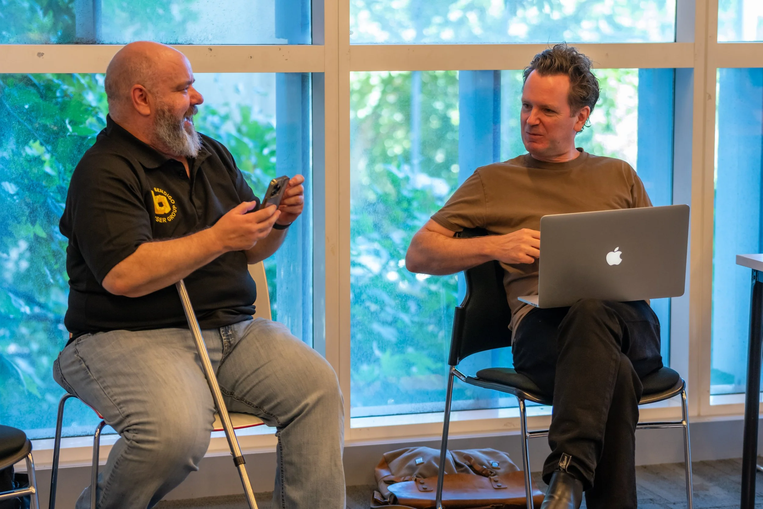 Two humans sit and converse. One has a walking stick, the other has a laptop.