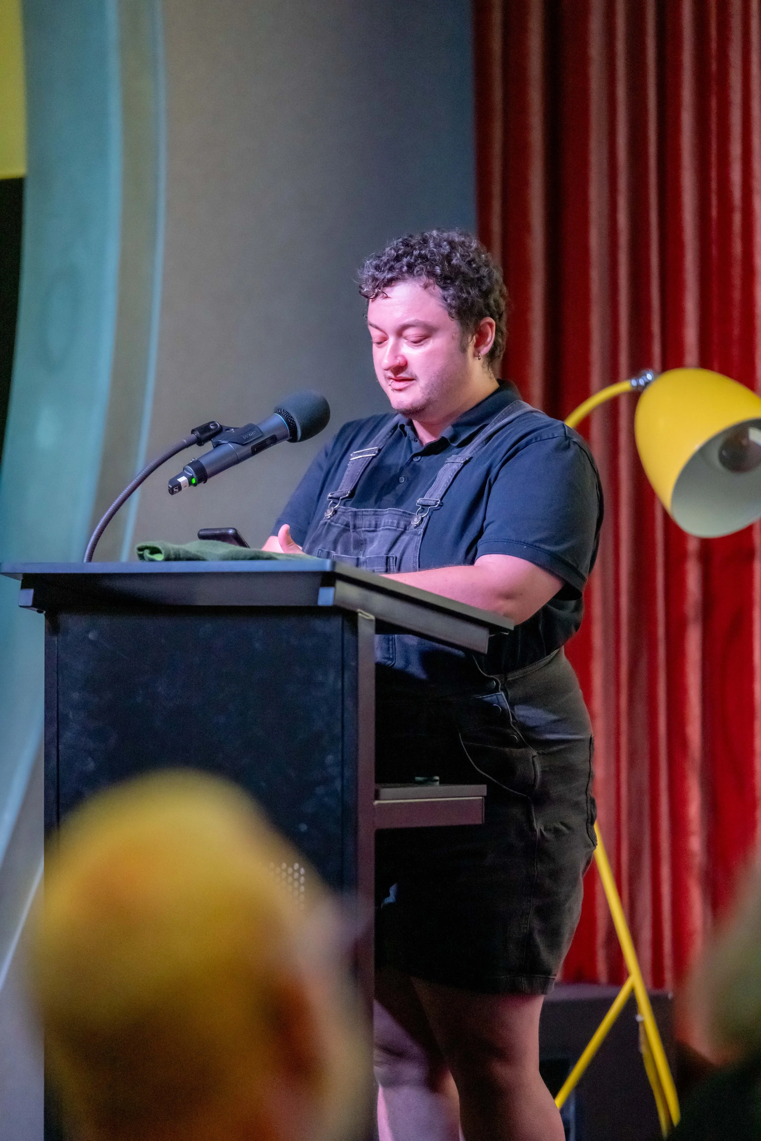 A human wearing black overalls speaks at a lecturn.