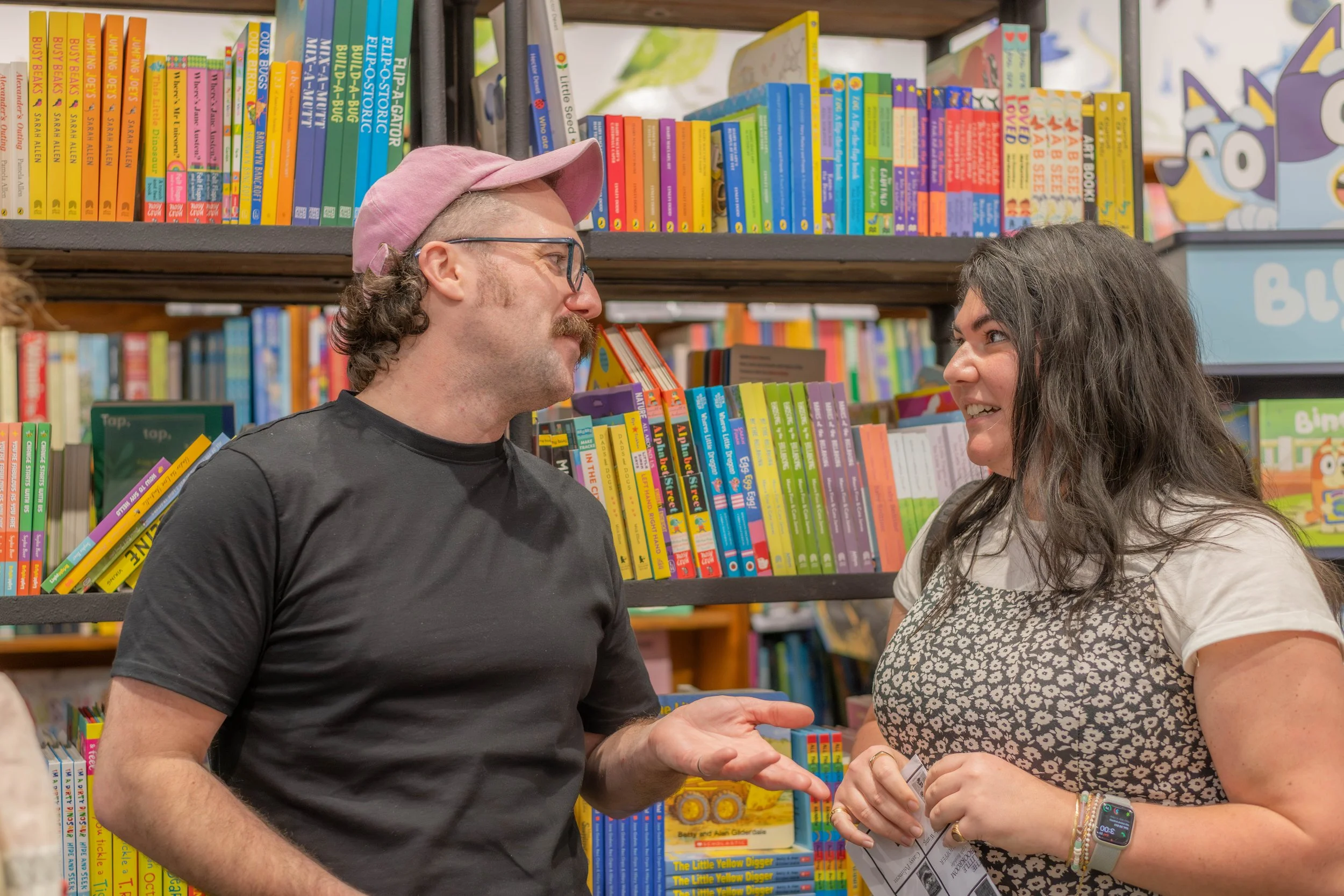 A human wearing a pink cap gesticulates whilst talking with a human in a dress.