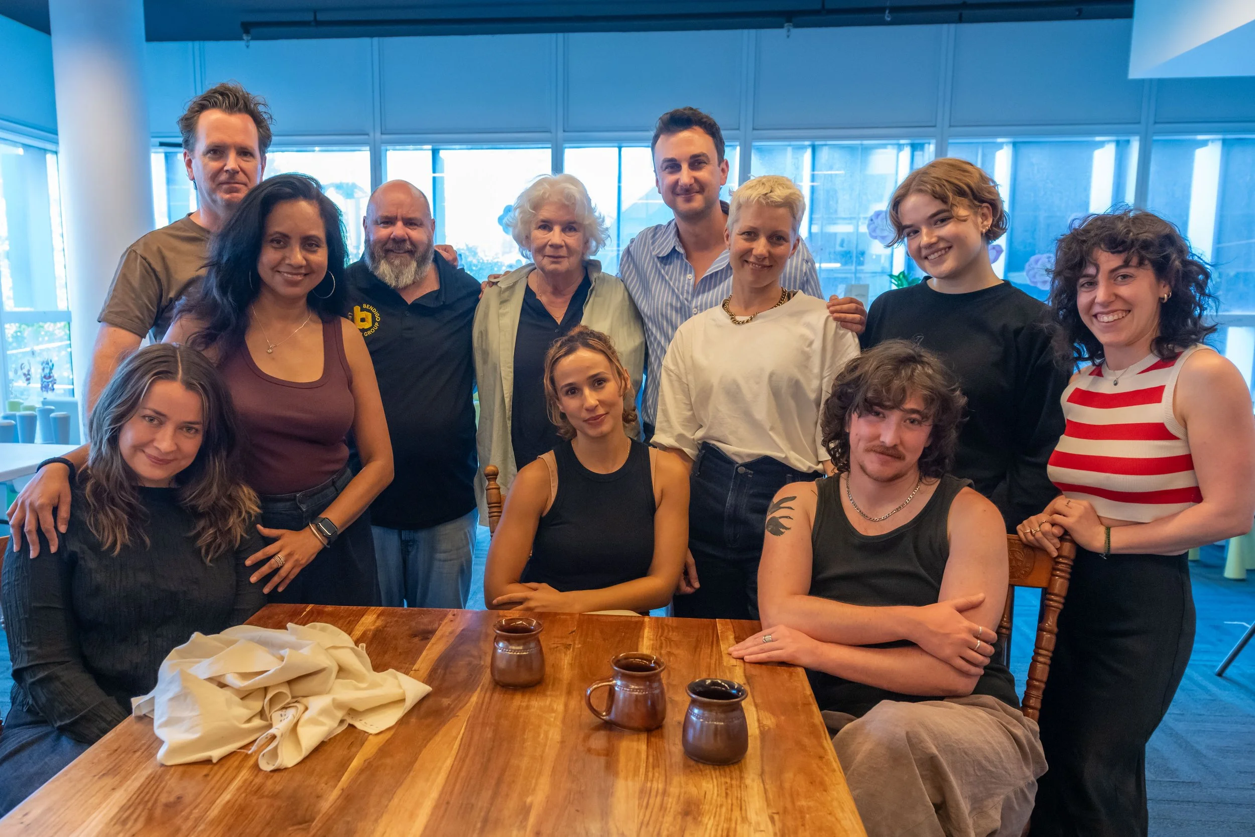 The cast and crew sit and stand around a brown coffee table, smiling at the camera.