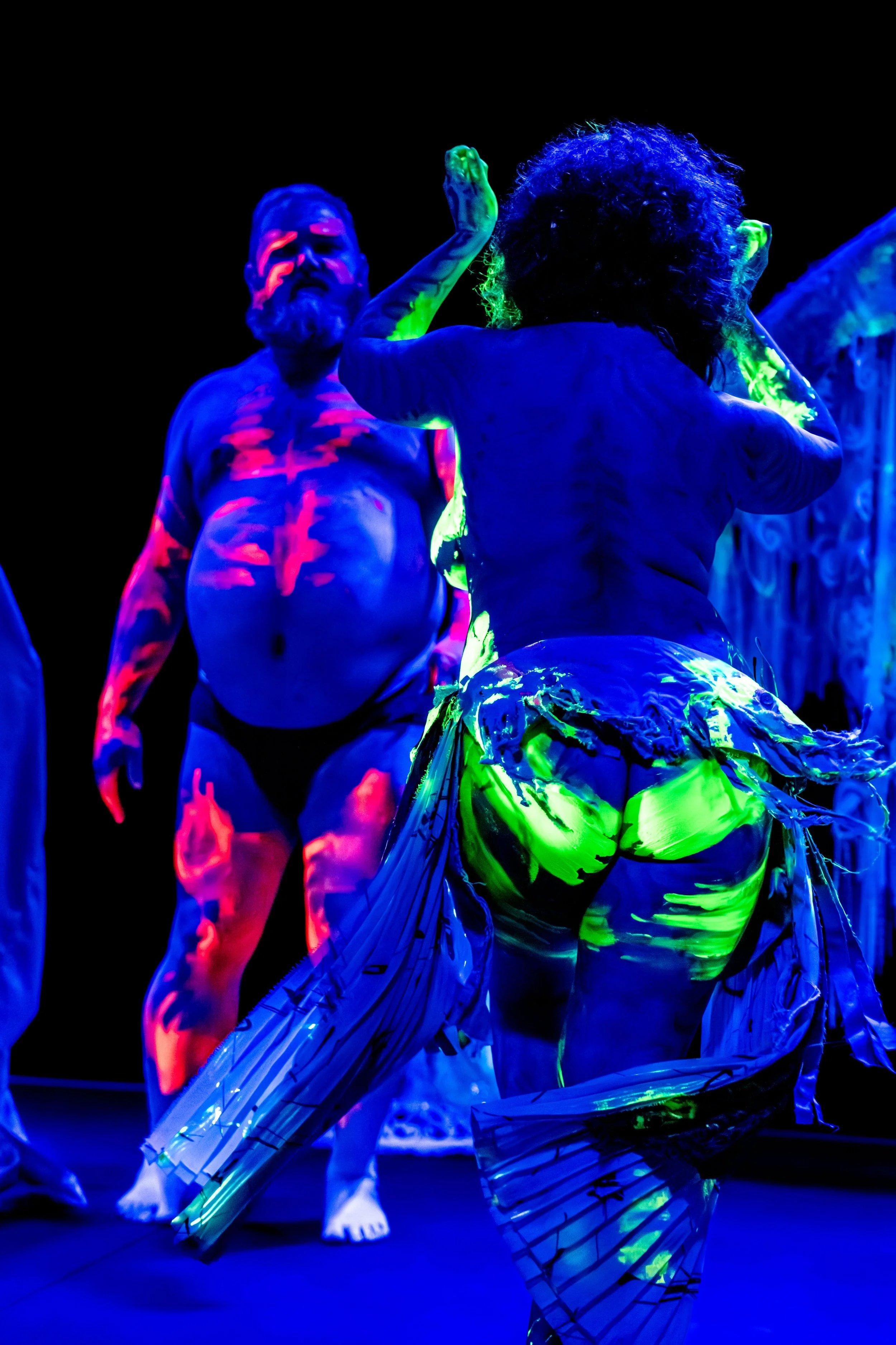 Yellow glow in the dark paint is smeared on the buttocks of a performer. A performer in the background has pink glow in the dark paint smeared across their body.