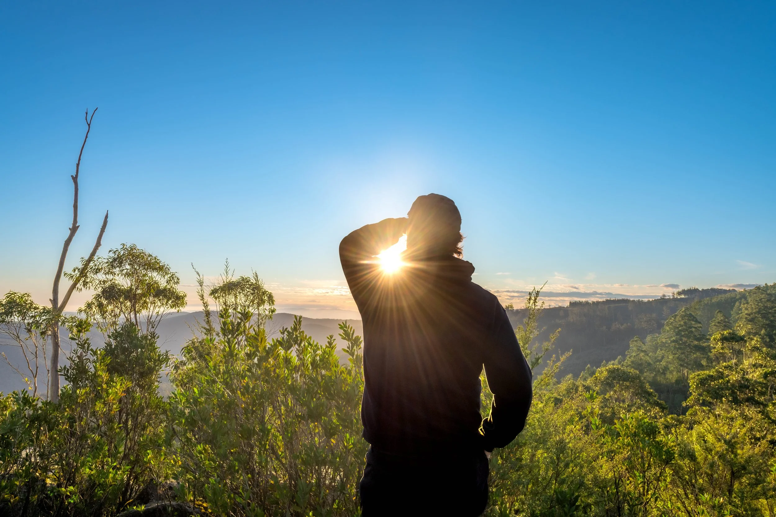 Silhouette of a human shielding their eyes from the setting sun as they view the mountainous vista beyond.