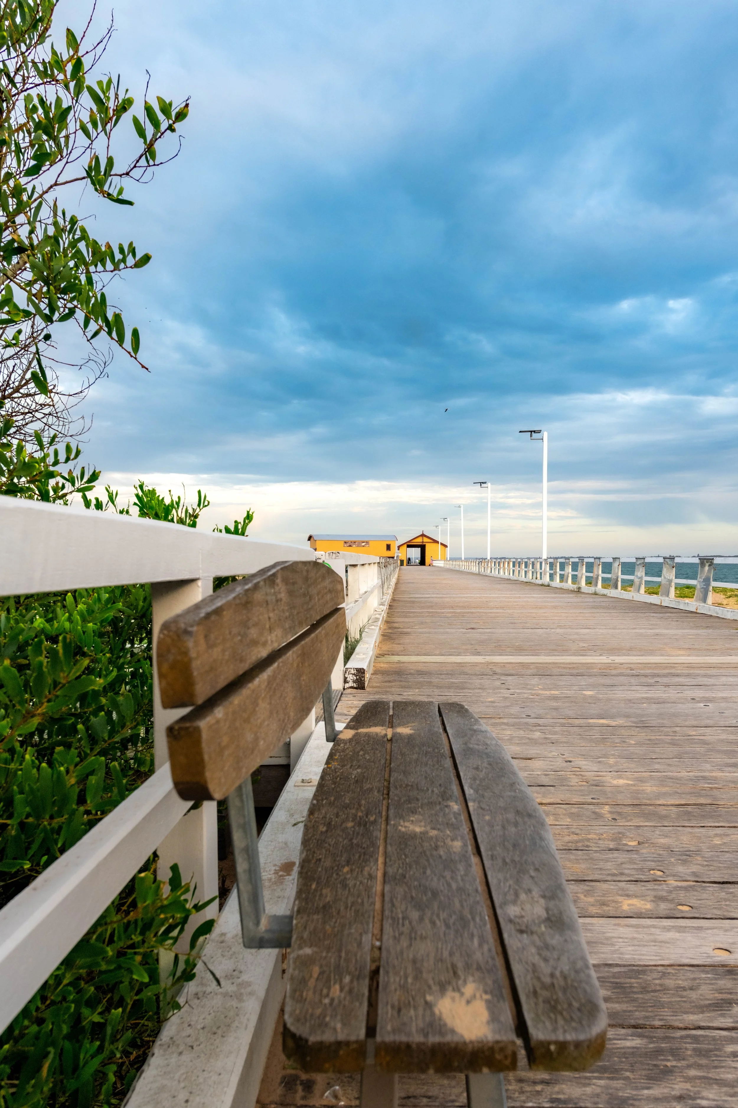 A wooden seat leads the eye to an orange building at the end of a white pier.