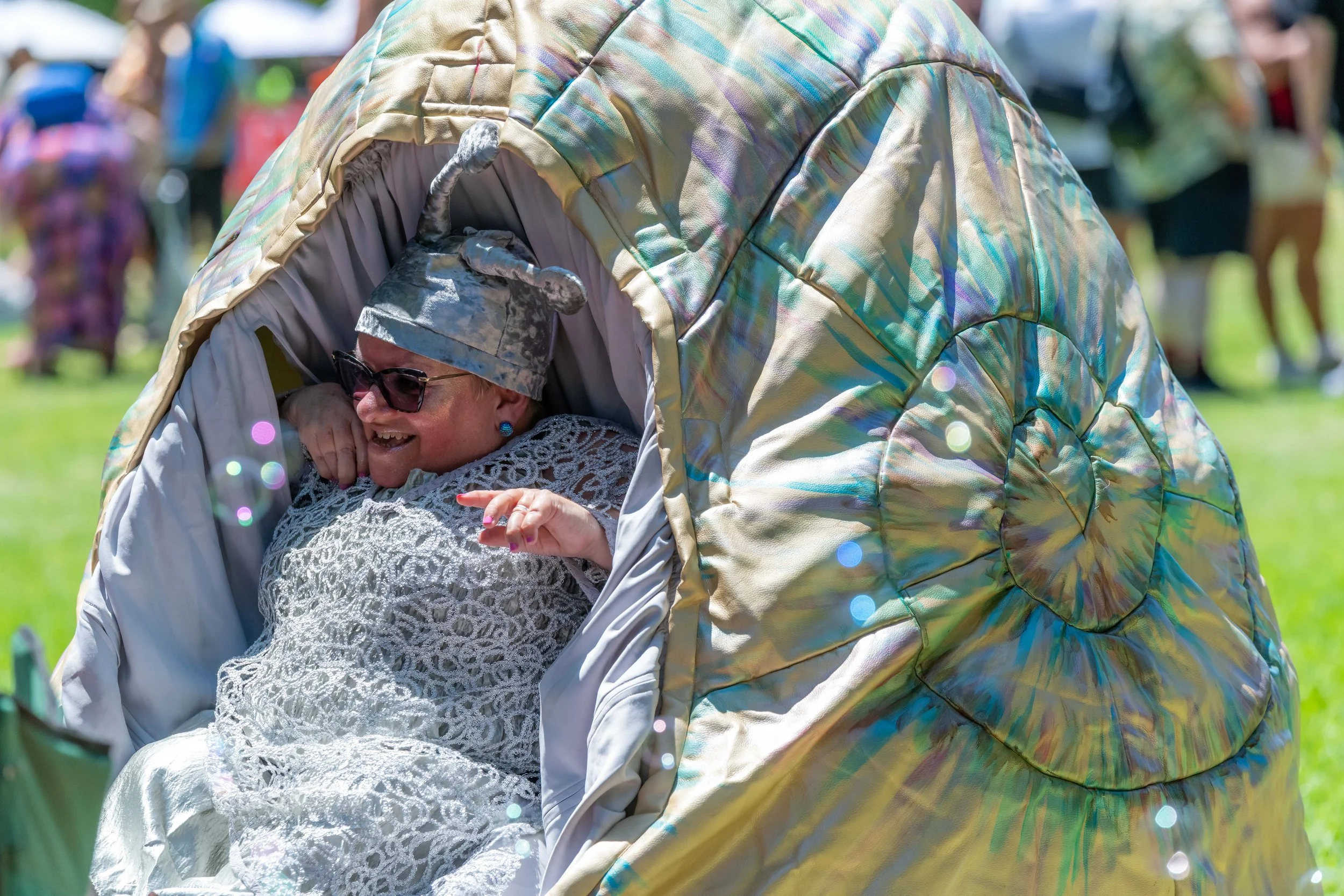 A small adult human is dressed in silver and wrapped in a large snail shell. Bubbles are floating by.