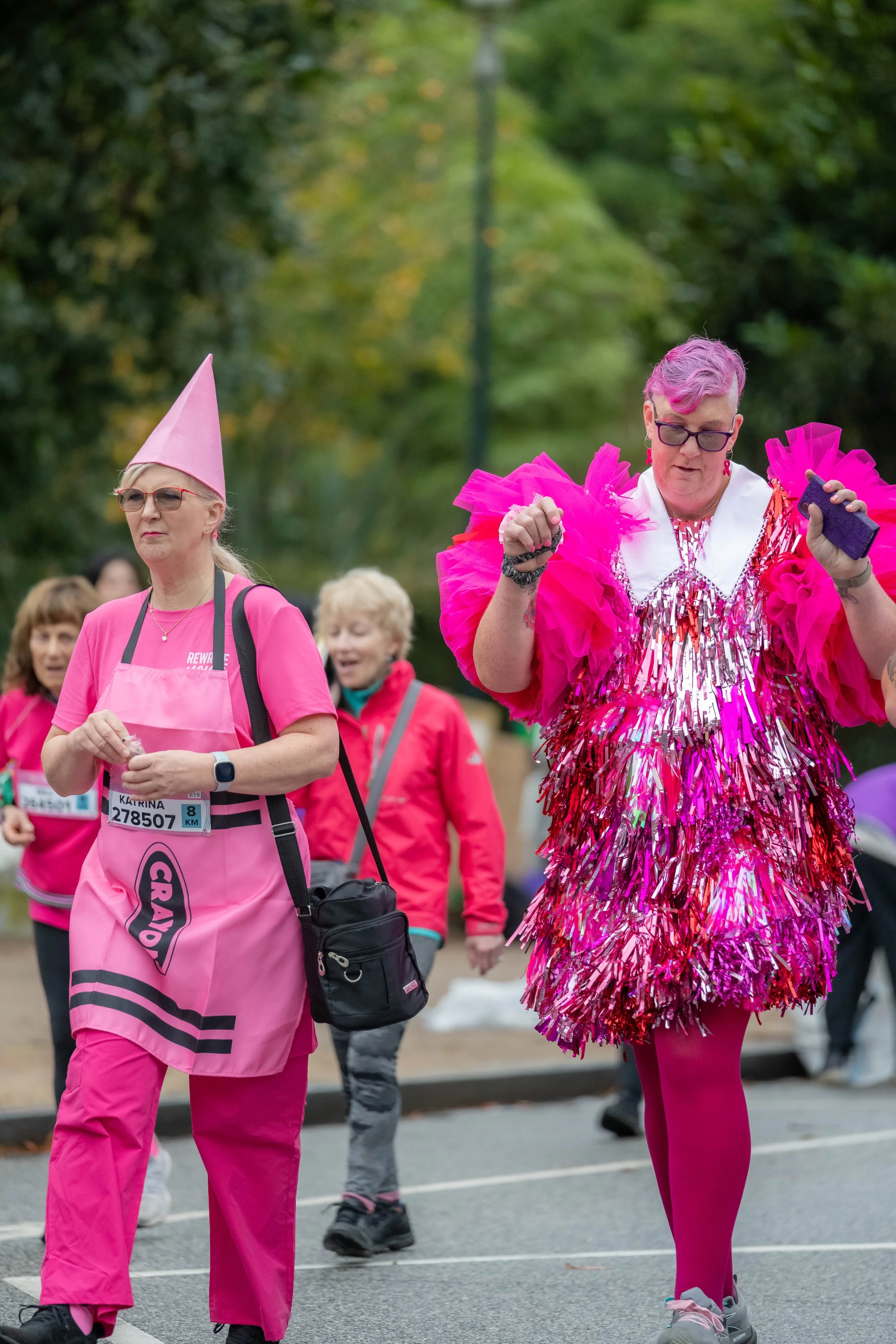 A racegoer is dressed as a pink crayon and walks next to a human in a pink shimmery outfit.