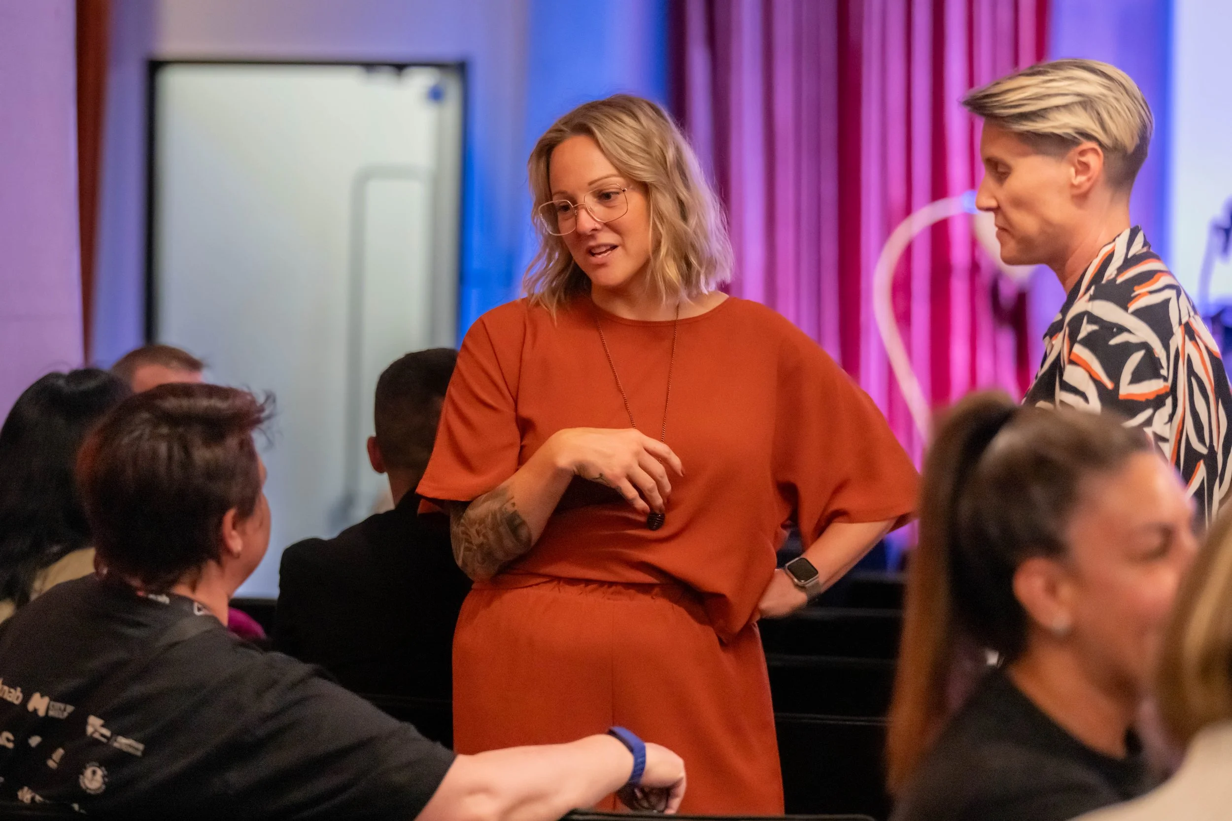 A human in a brown suit stands and chats with a human sitting down.
