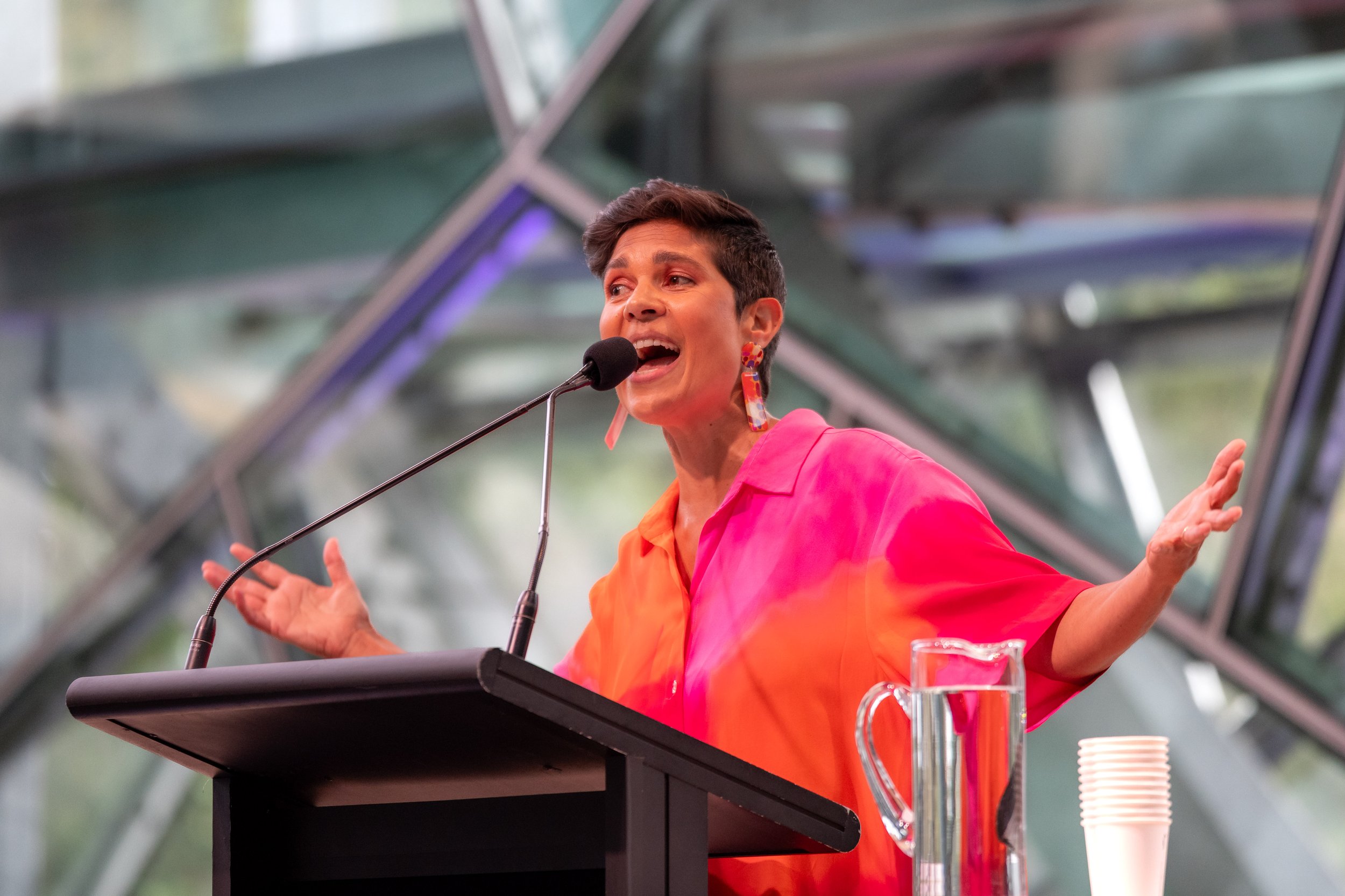 Narelda Jacobs speaks into a microphone, their arms are stretched outward.