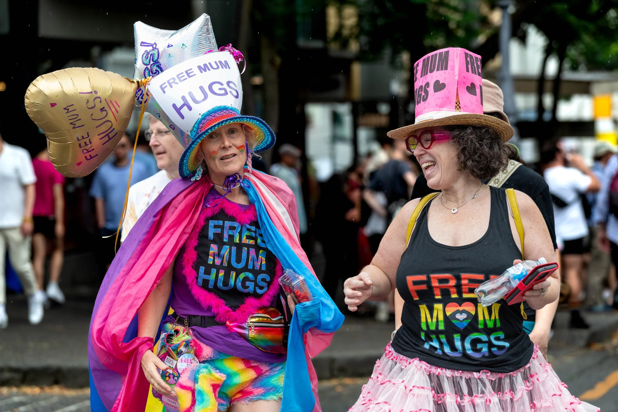 Two humans wear hats and singlets that state "Free mum hugs".