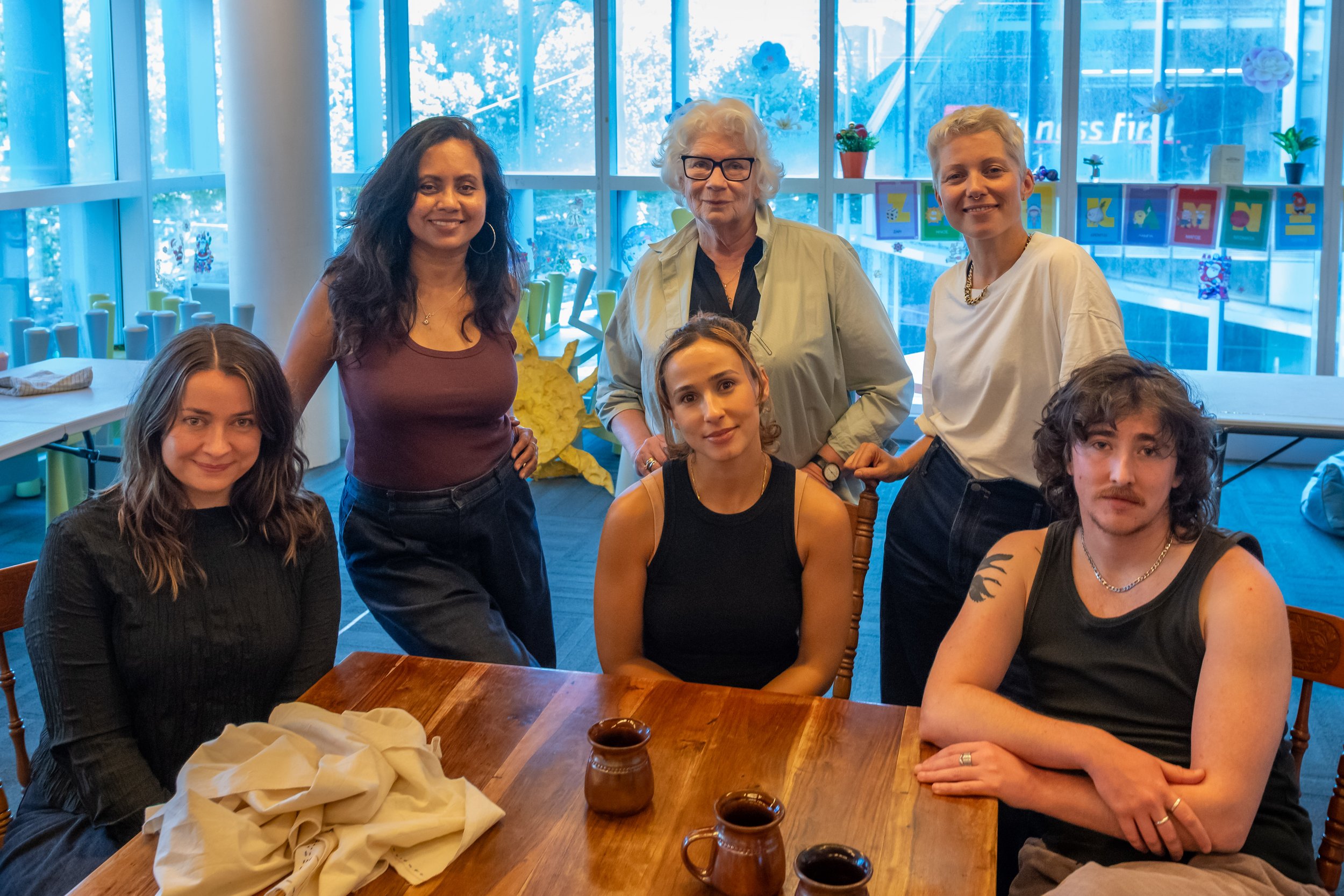 The six actors of the play sit and stand around a brown coffee table.