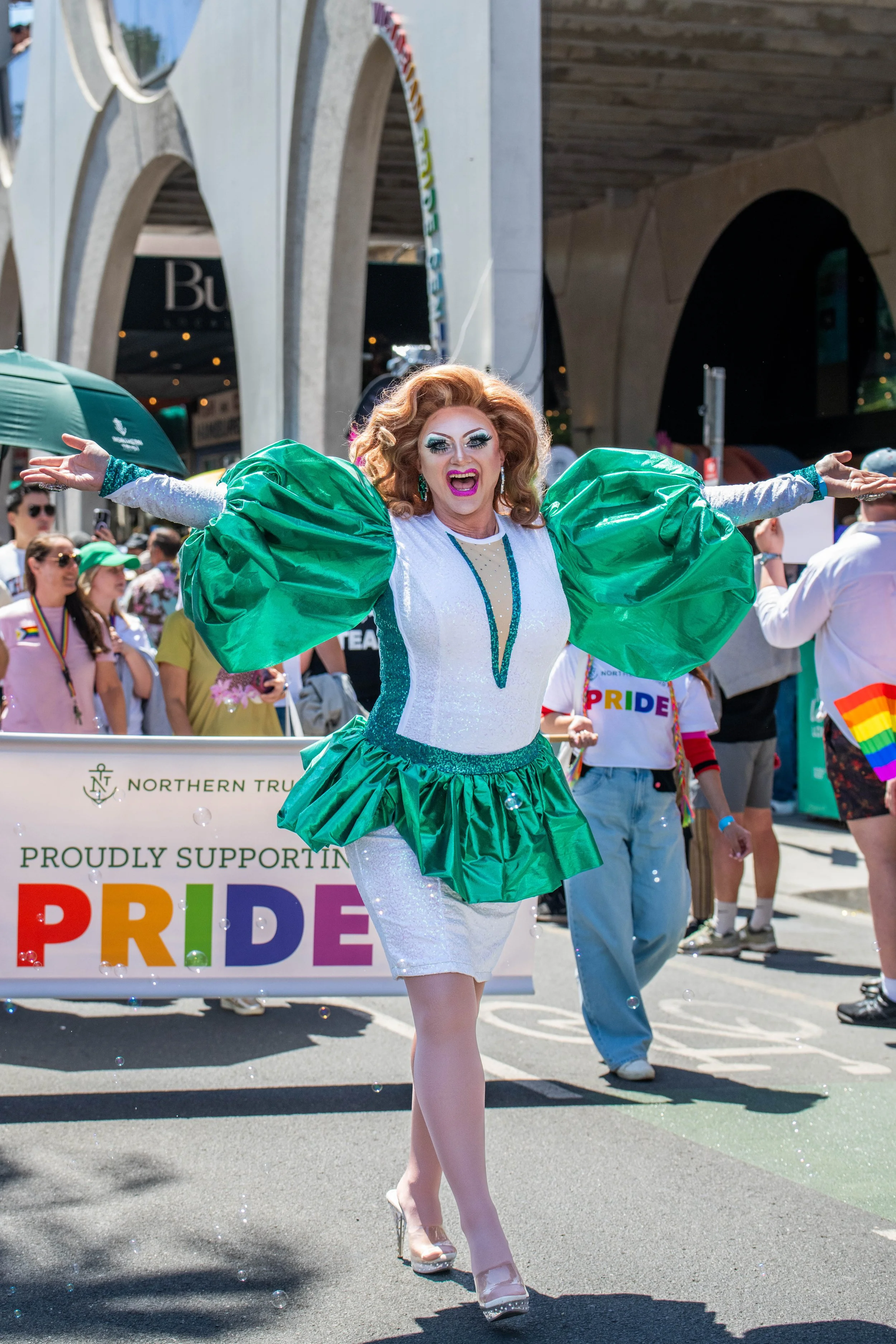 A drag queen outstretches her arms and smiles open-mouthed at the camera.