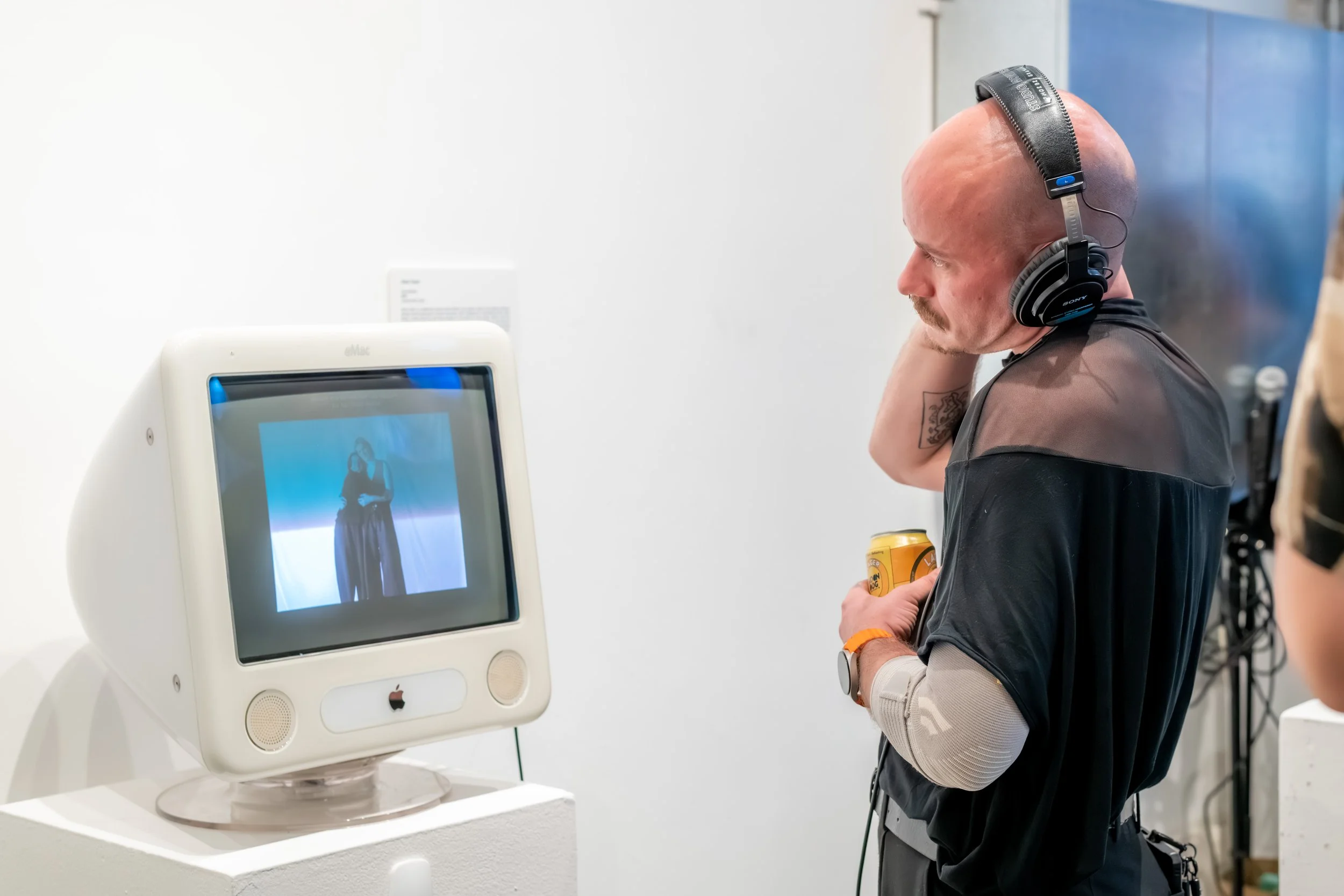 A human looks at an old mac computer. They are listening to the artwork through headphones.