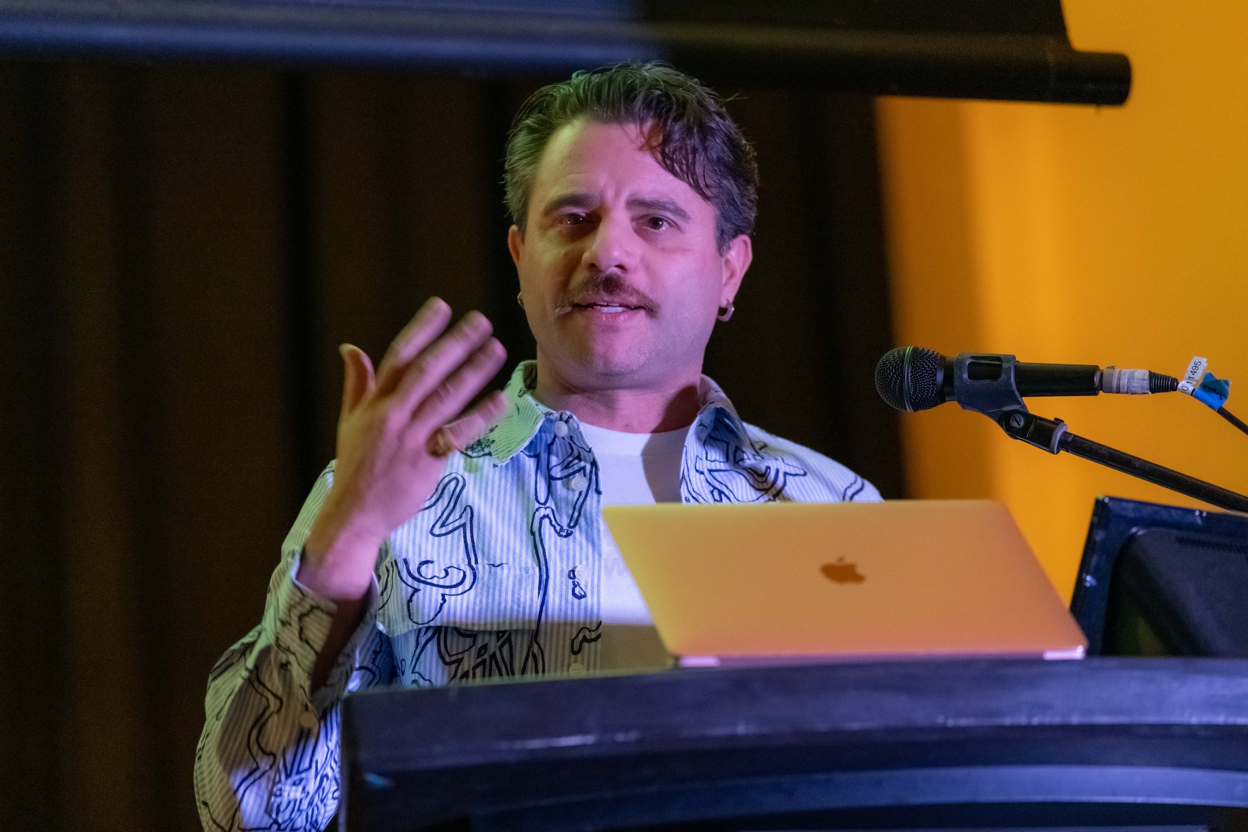 Sam Stiegler stands behind a lecturn and speaks into a microphone.