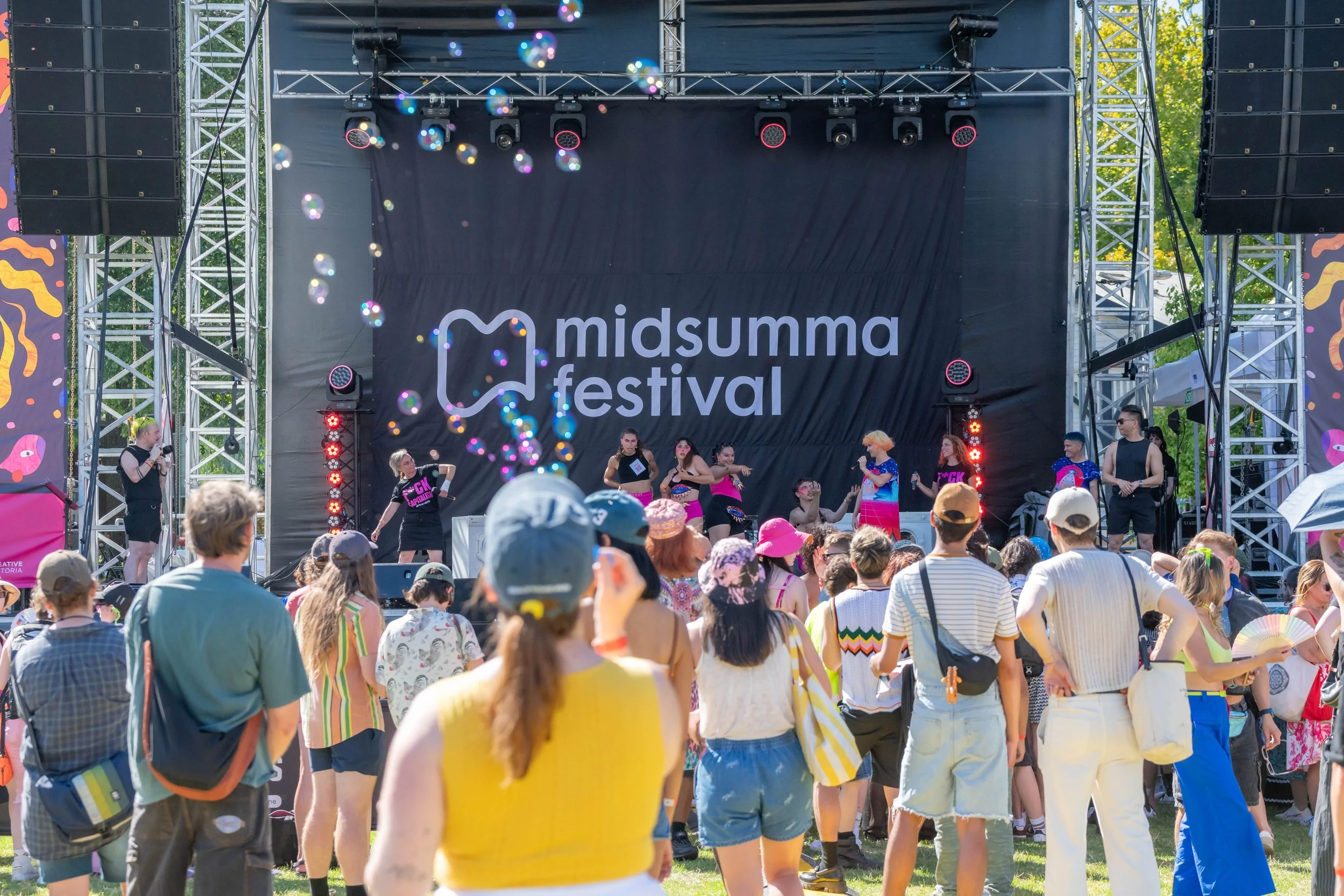 A human in a yellow top watches the Midsumma Carnival stage and blows bubbles into the air.