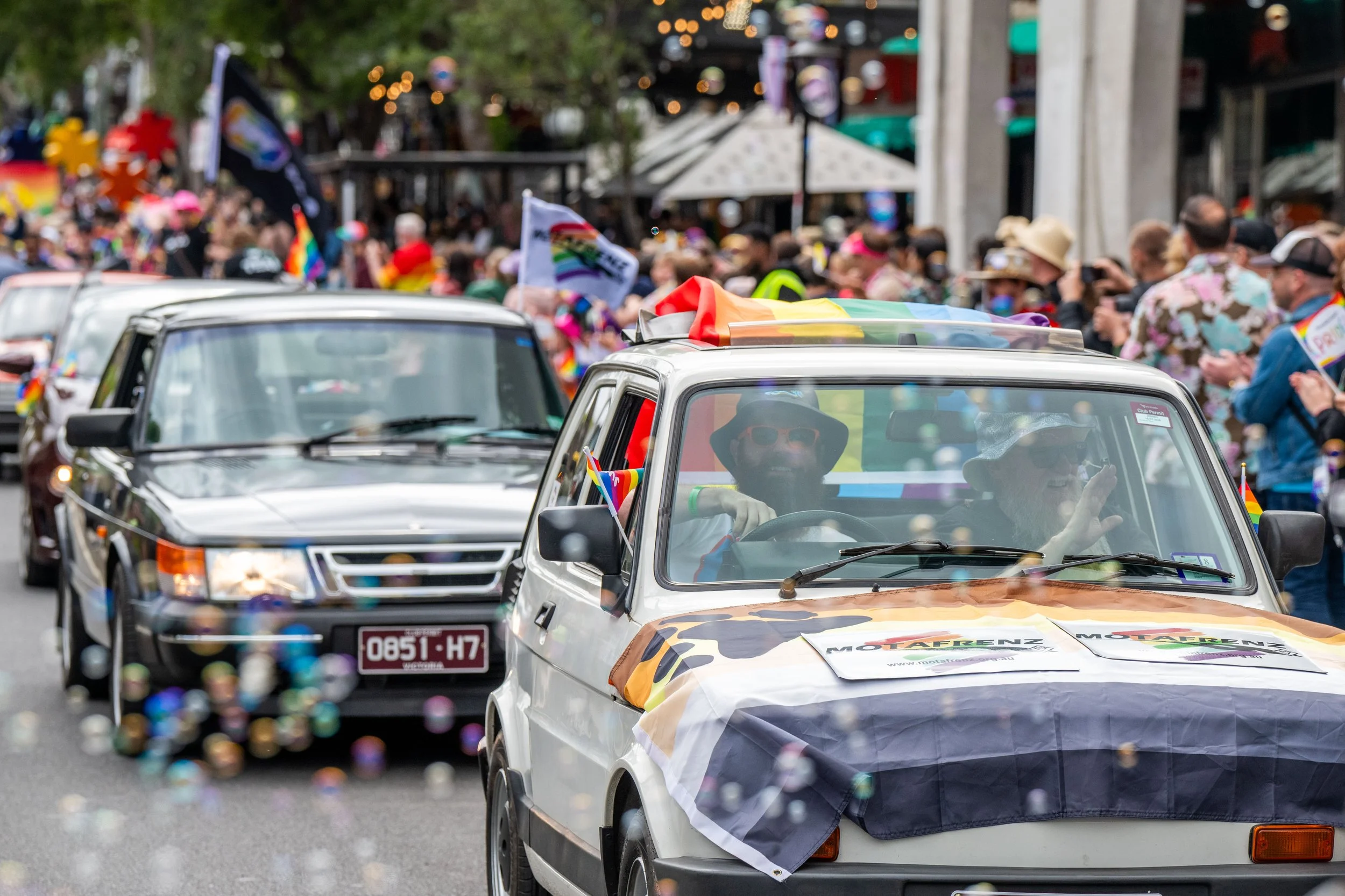 Bubbles blowing across vintage cars. The leading car has a bear pride flag on the bonnet.