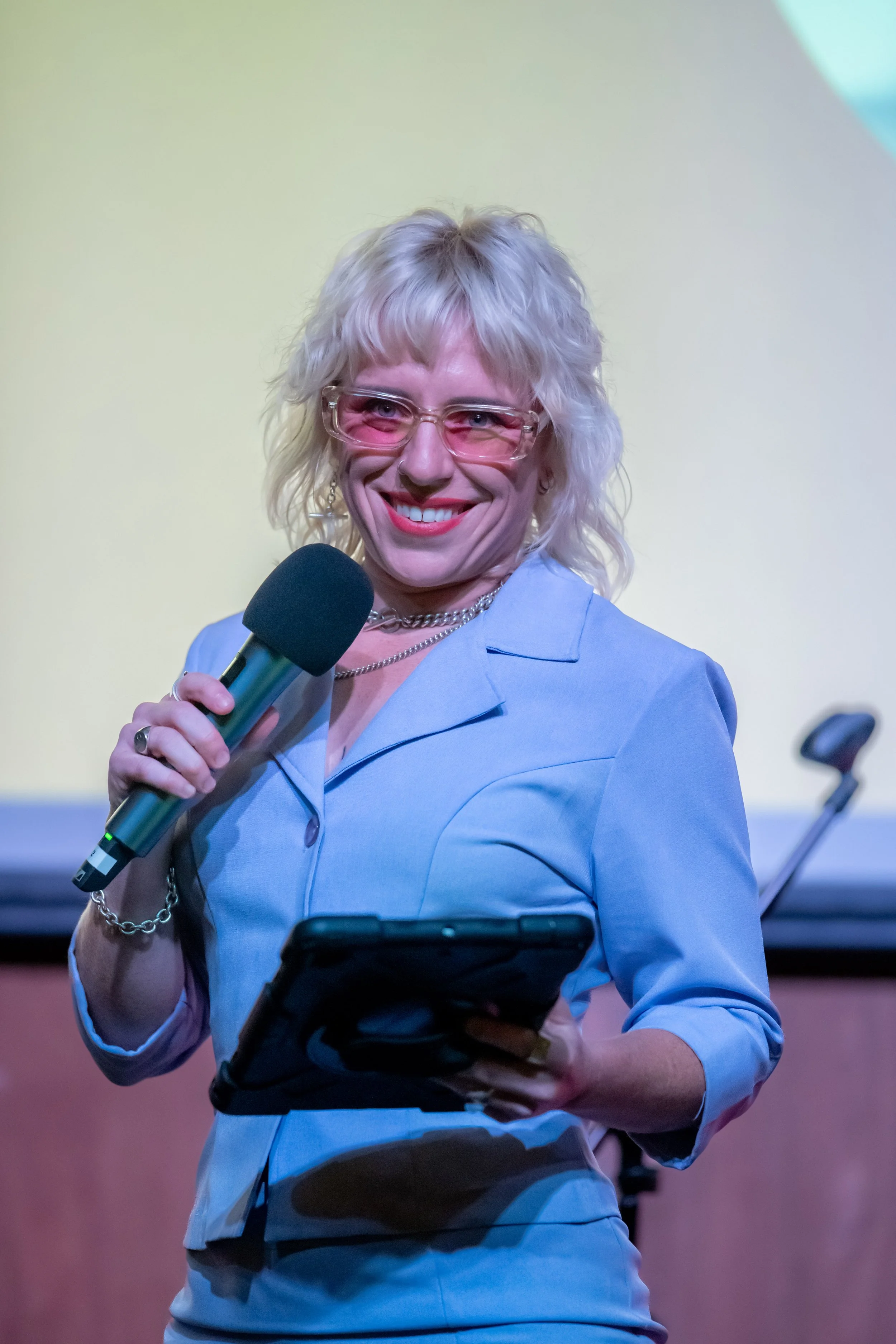 A human in a blue suit and pink glasses smiles as they talk into a microphone.