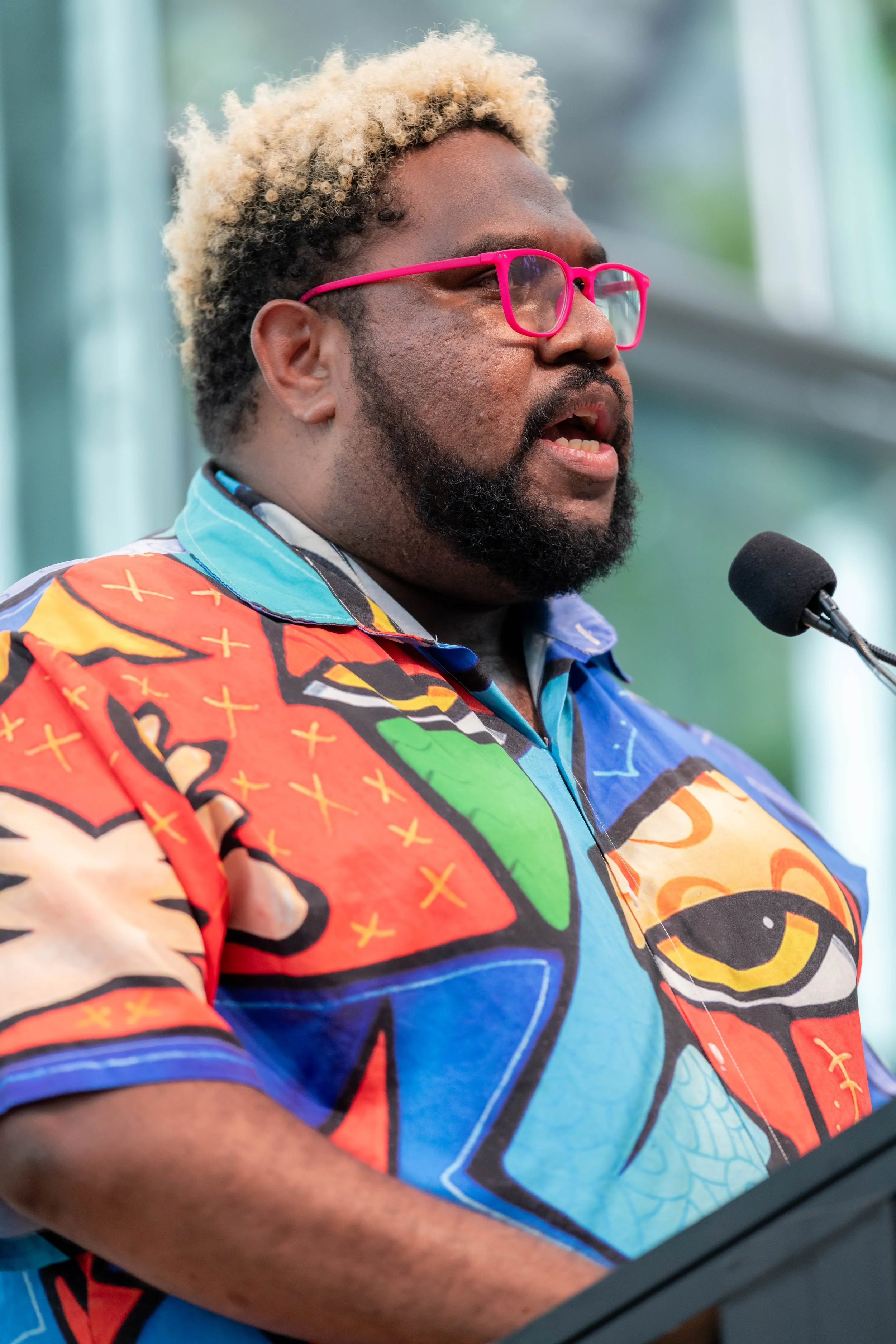 Jay Wymarra wears pink glasses and speaks into a microphone.