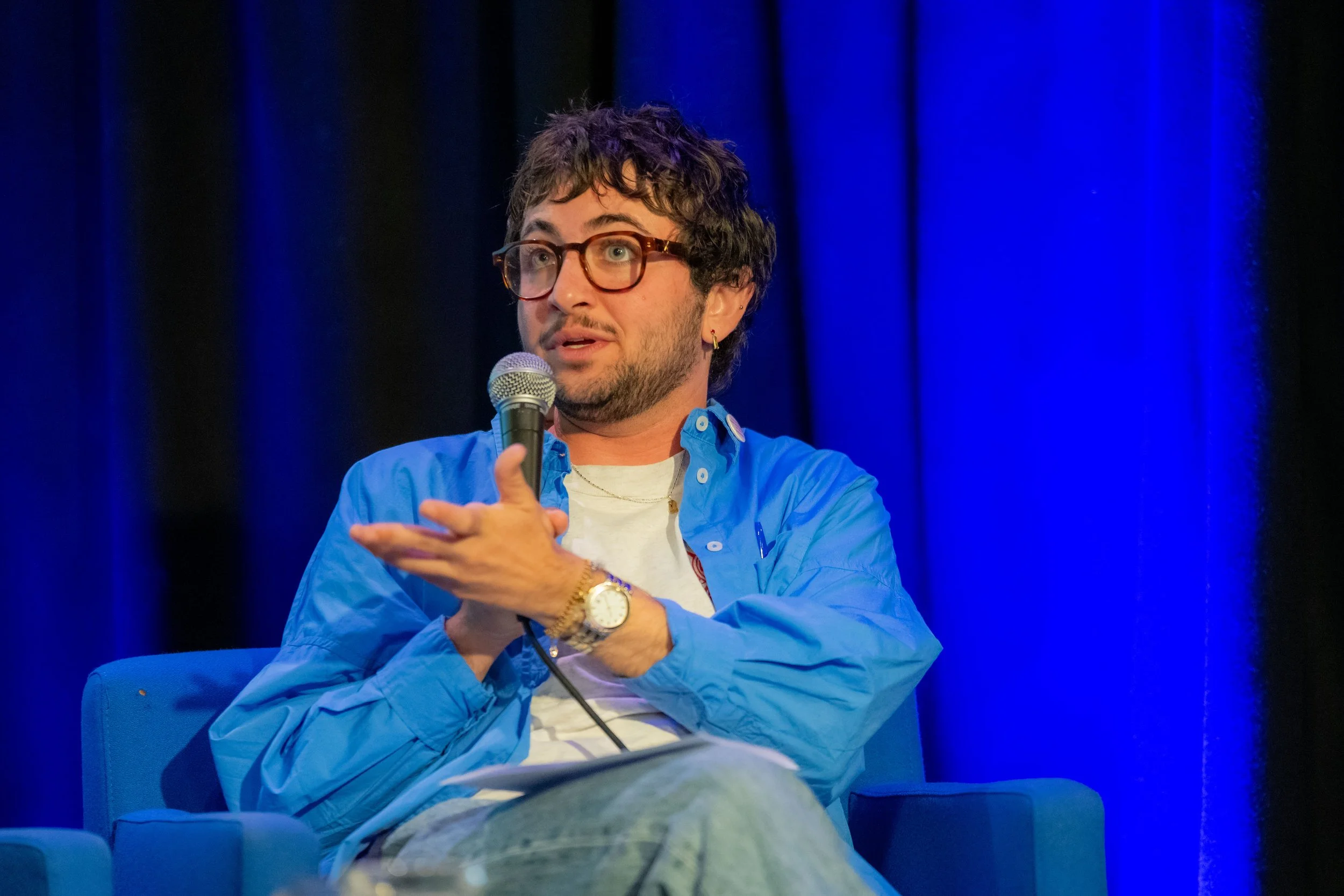 A panelist in a blue shirt speaks into a microphone.
