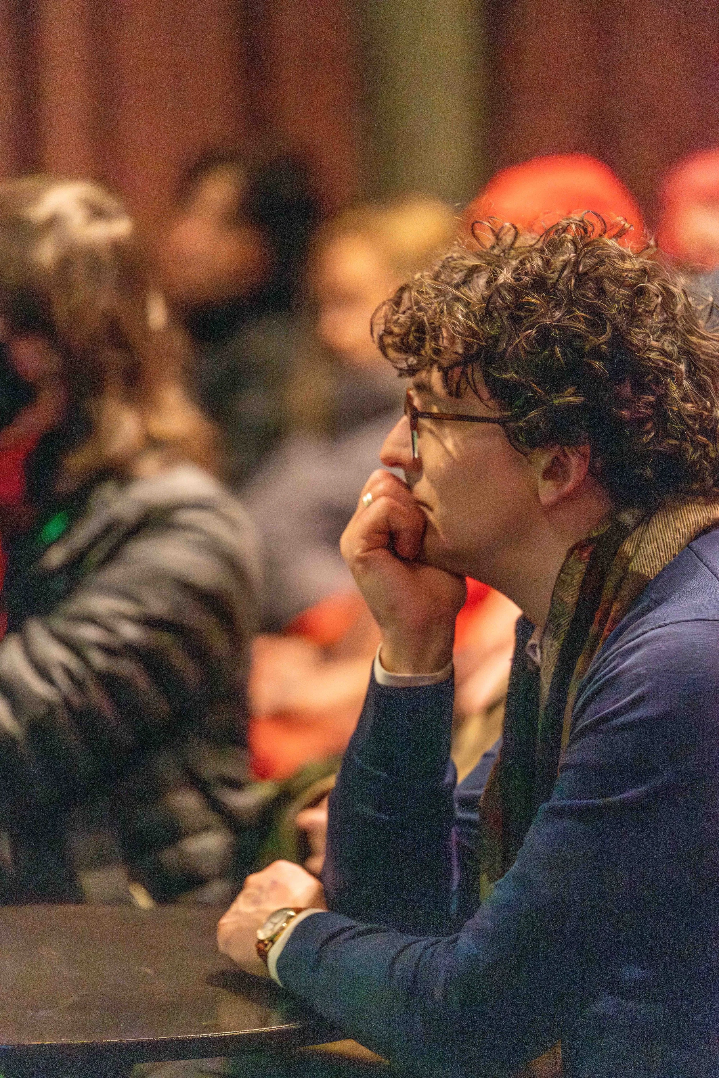A human holds their hand to their mouth and leans slightly forward in thouoght as they listen to the unseen panelists.